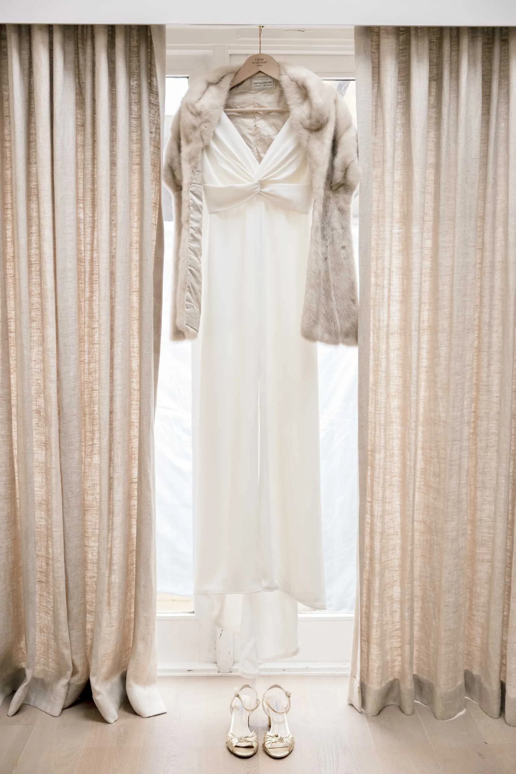 Wedding dress hanging in front of a window between beige curtains with a pair of white shoes on the floor beneath.