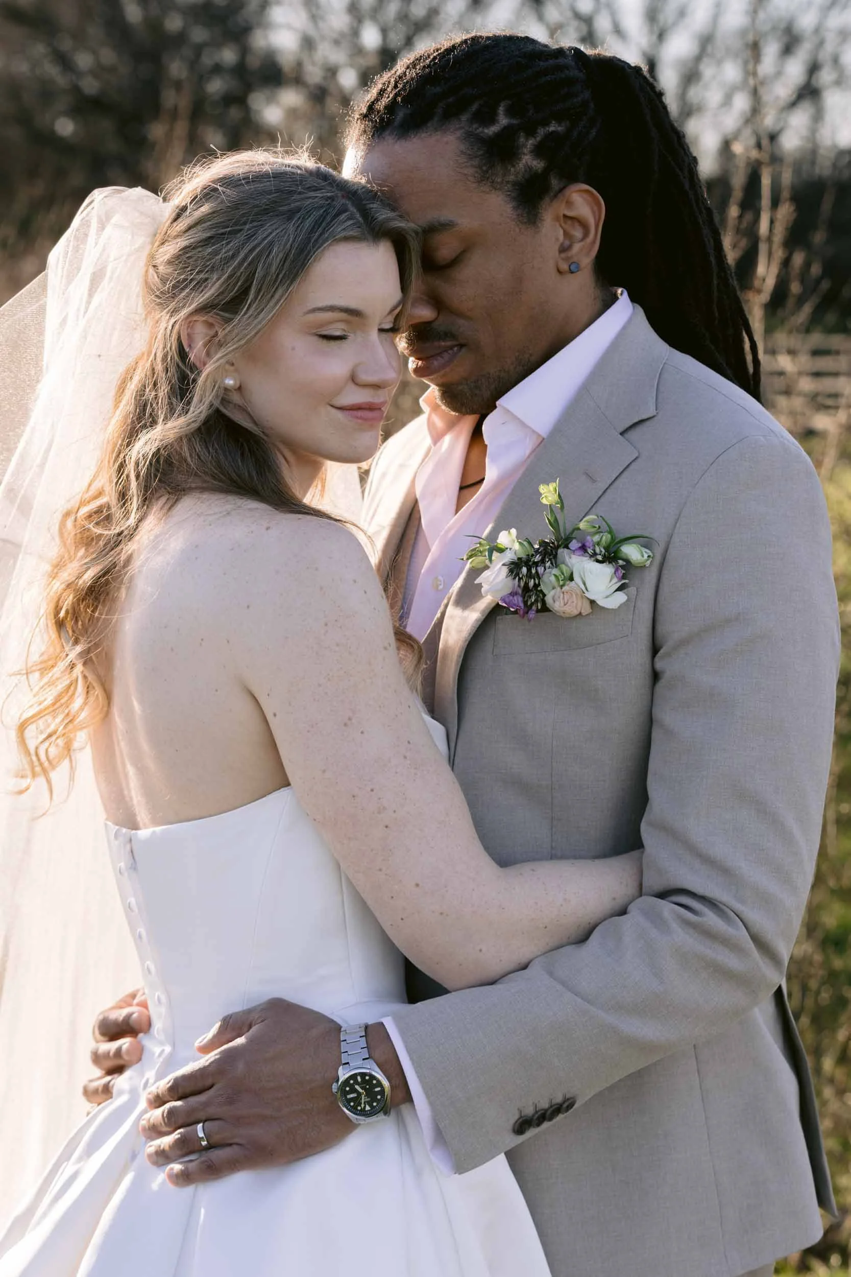 A bride and groom embracing outdoors during sunset, with eyes closed and foreheads touching, smiling gently.