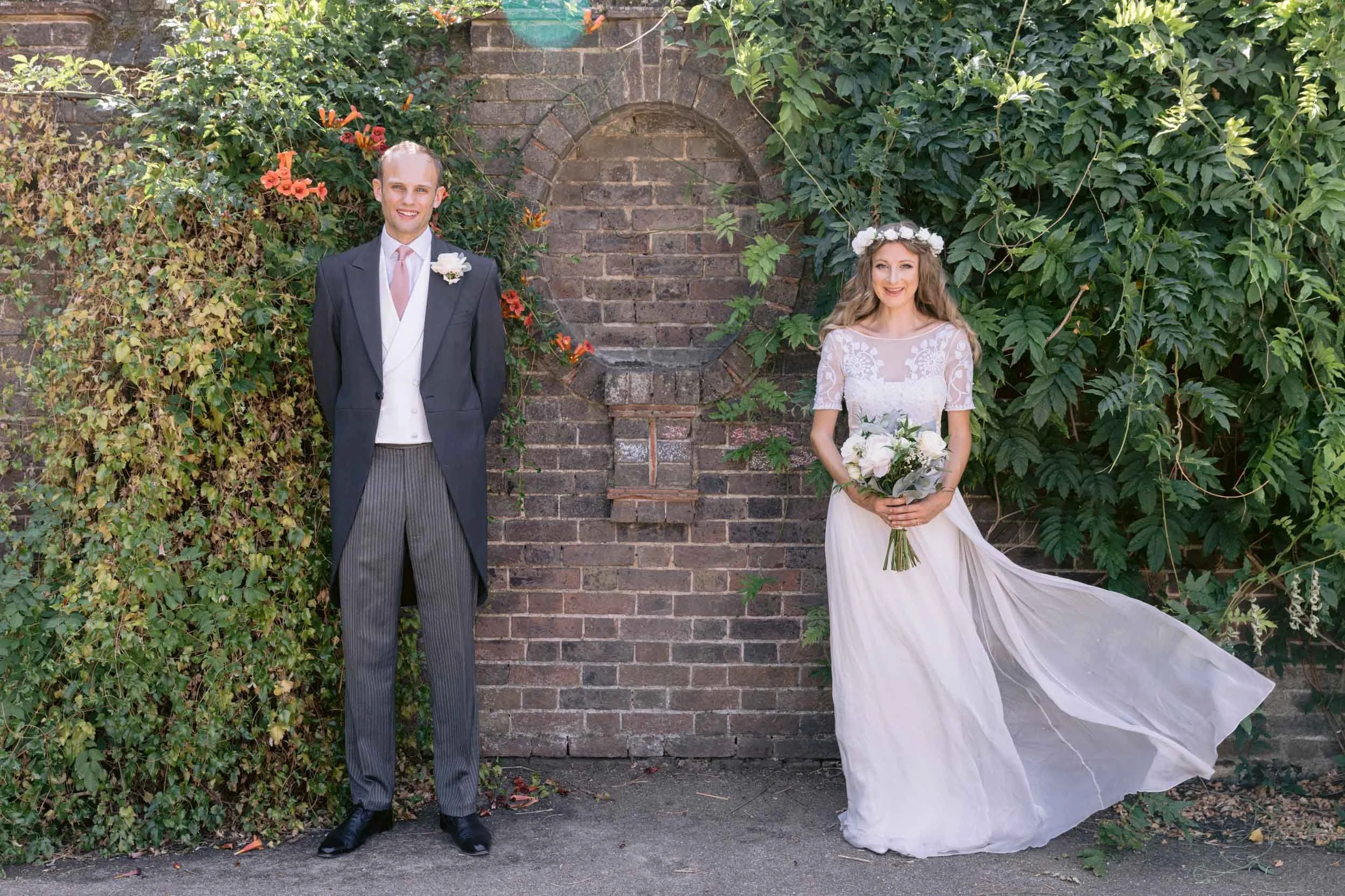 A bride and groom standing outdoors in front of a brick wall covered with green foliage and orange flowers. The groom is wearing a dark suit with a pink tie and white boutonniere. The bride is wearing a white lace wedding dress with sheer sleeves, a floral crown, and is holding a bouquet of white roses. The bride's dress has a long train flowing to the ground.