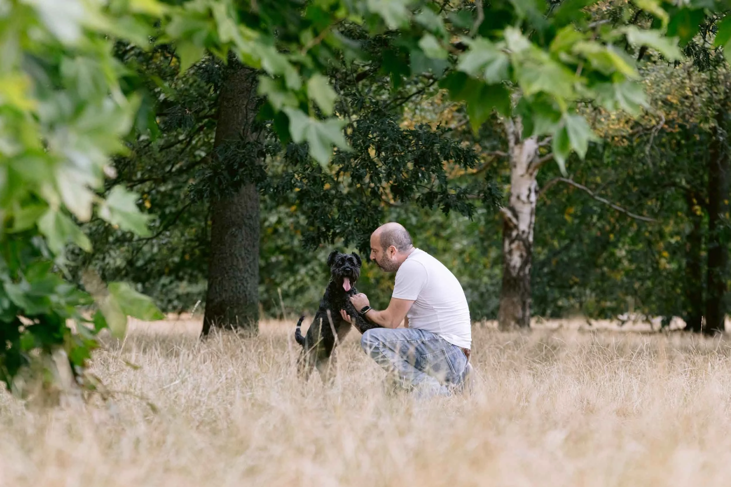 A man kneeling on the grass holding a black dog with a white chest outdoors in a wooded area.