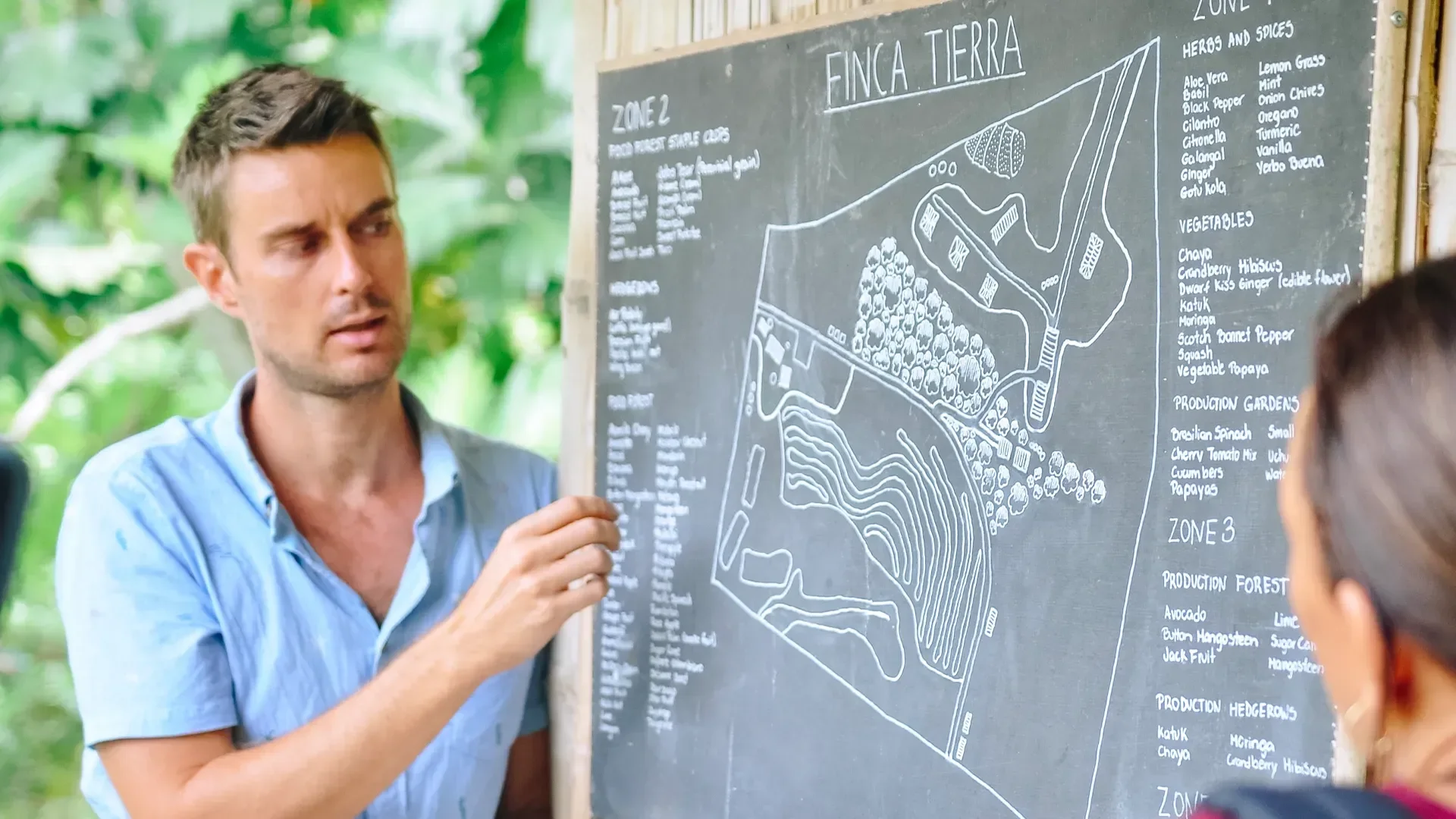 Instructor explaining a tropical permaculture site design on a hand-drawn master plan, showing garden zones, paths, and water systems