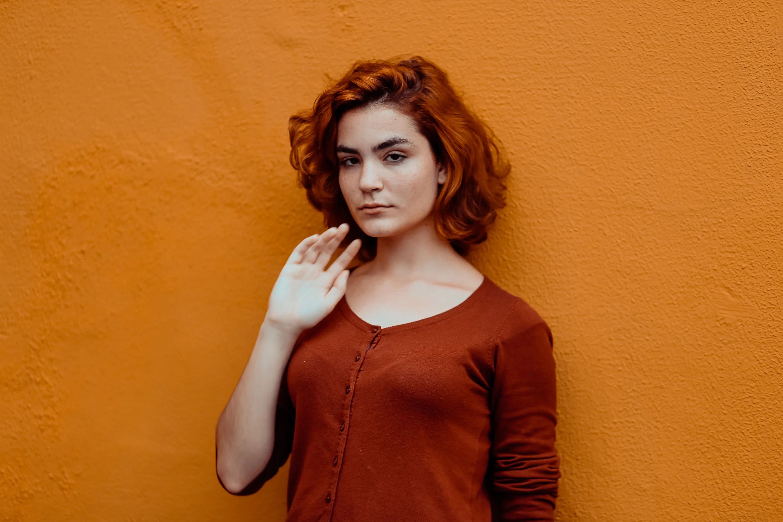 Image of unsmiling person with shoulder-length red hair standing next to an orange wall, via Unsplash