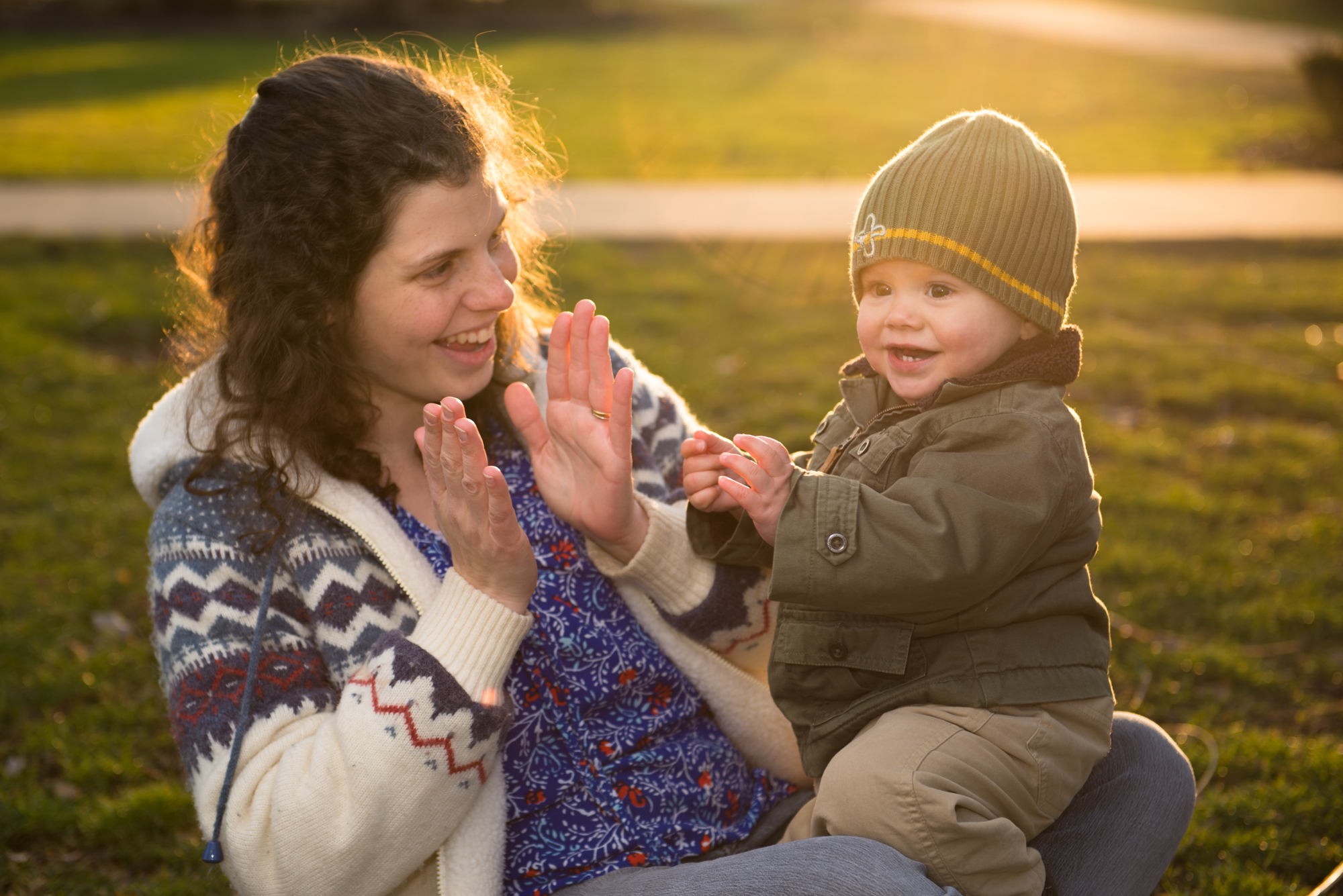 A mom sits in the park showing her young son to clap during a family photo shoot