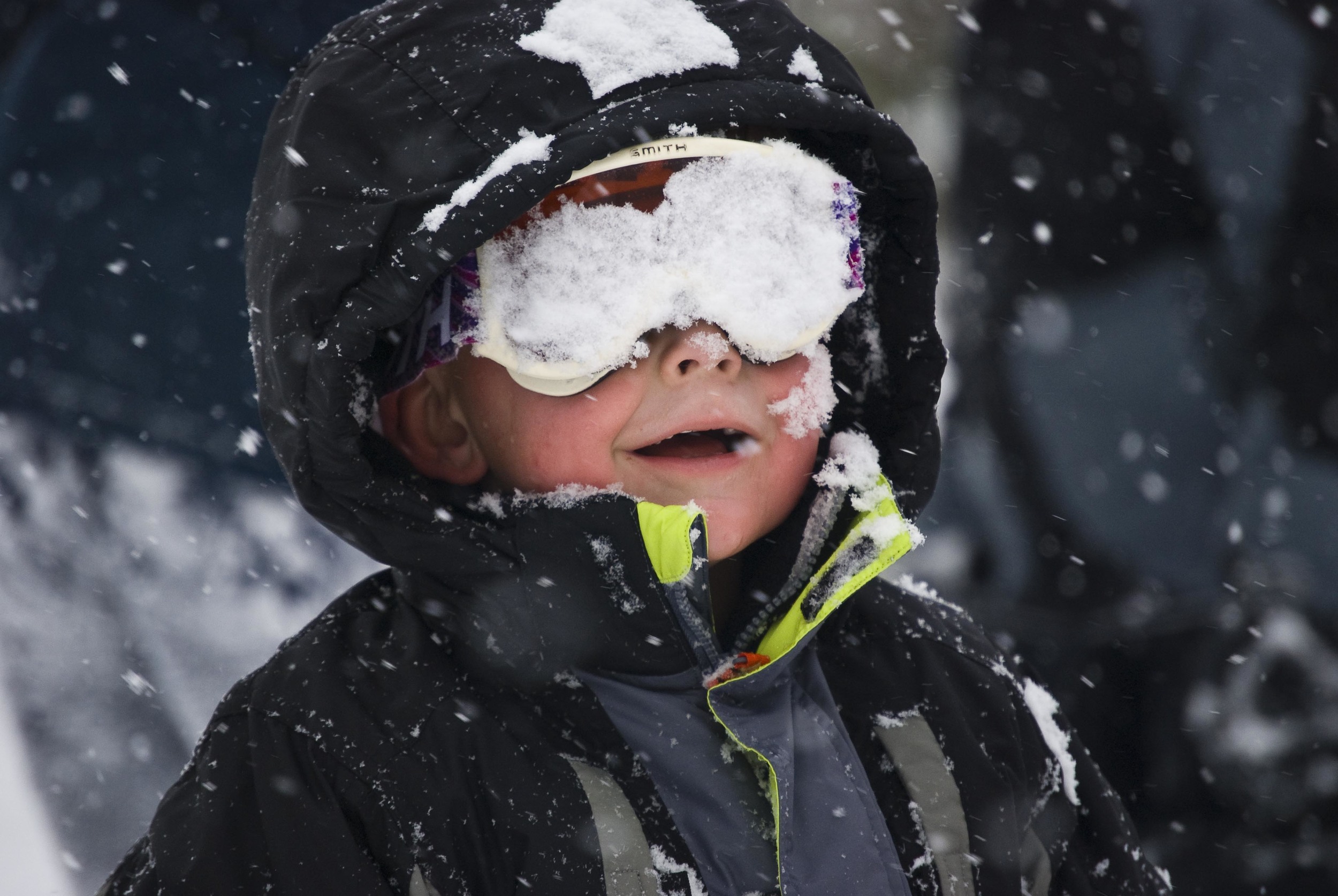 A toddler in snow gear with his googles caked with snow and a big grin