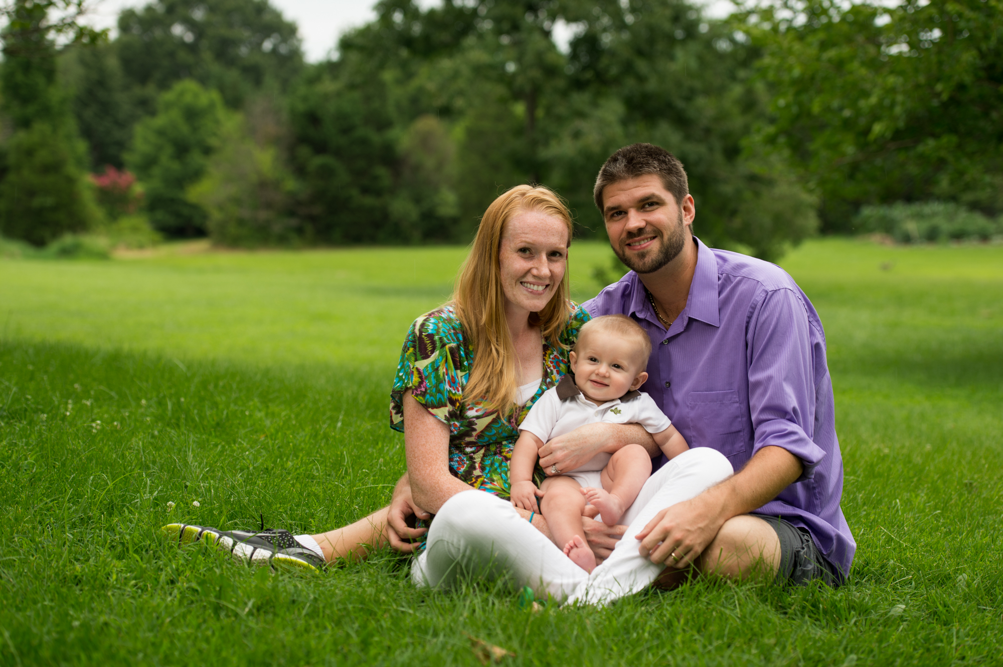 A young couple sits in a lush green field holding their baby boy for their family portrait