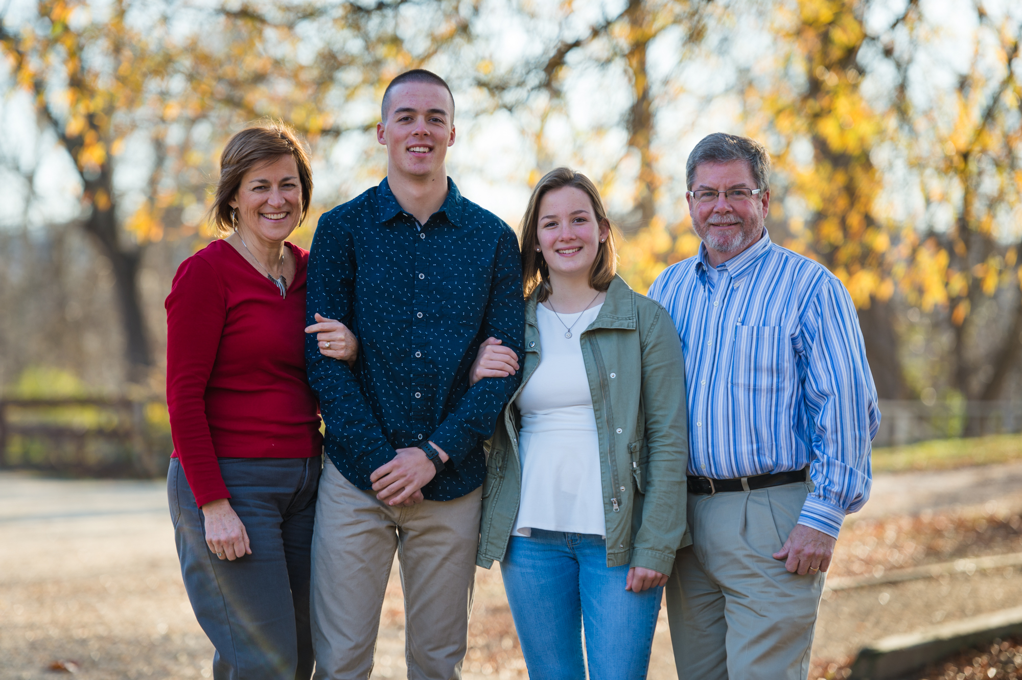A husband and wife stand arm in arm with their grown children amidst a beautiful fall backdrop for a family photo