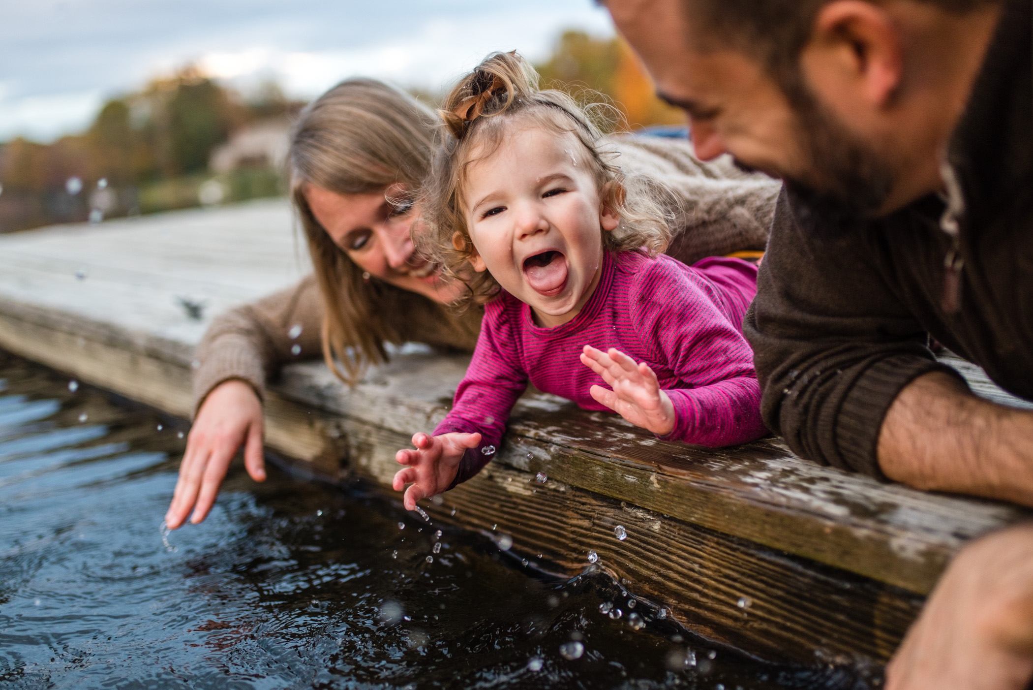 A toddler and her parents lay on a deck, splashing lake water and laughing.