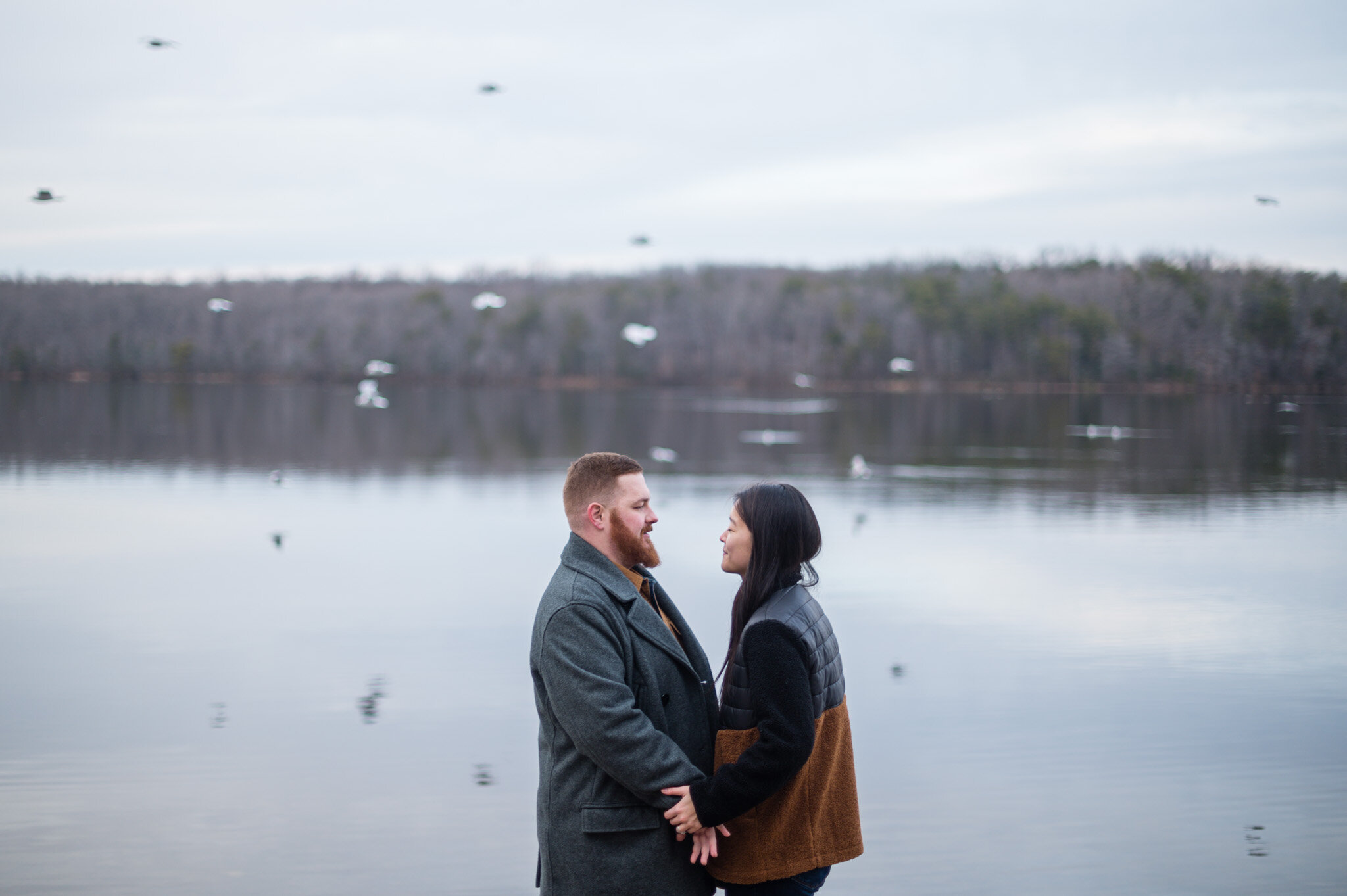 A couple stands in front of Burke Lake while a flock of birds flies behind them