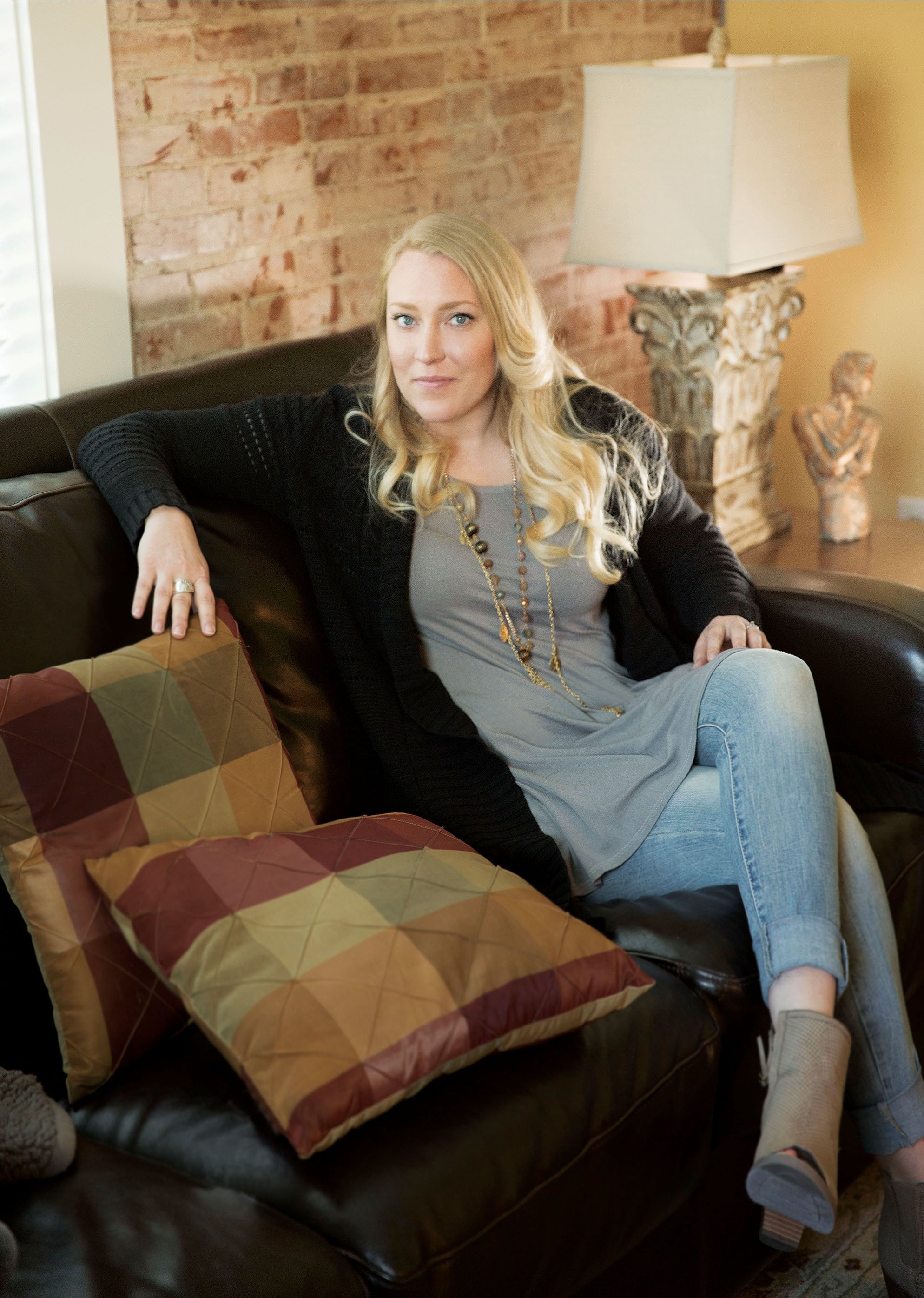 A blonde woman sitting on a black leather couch with patterned pillows in a living room with a brick wall, a decorative lamp, and sculptures in the background.