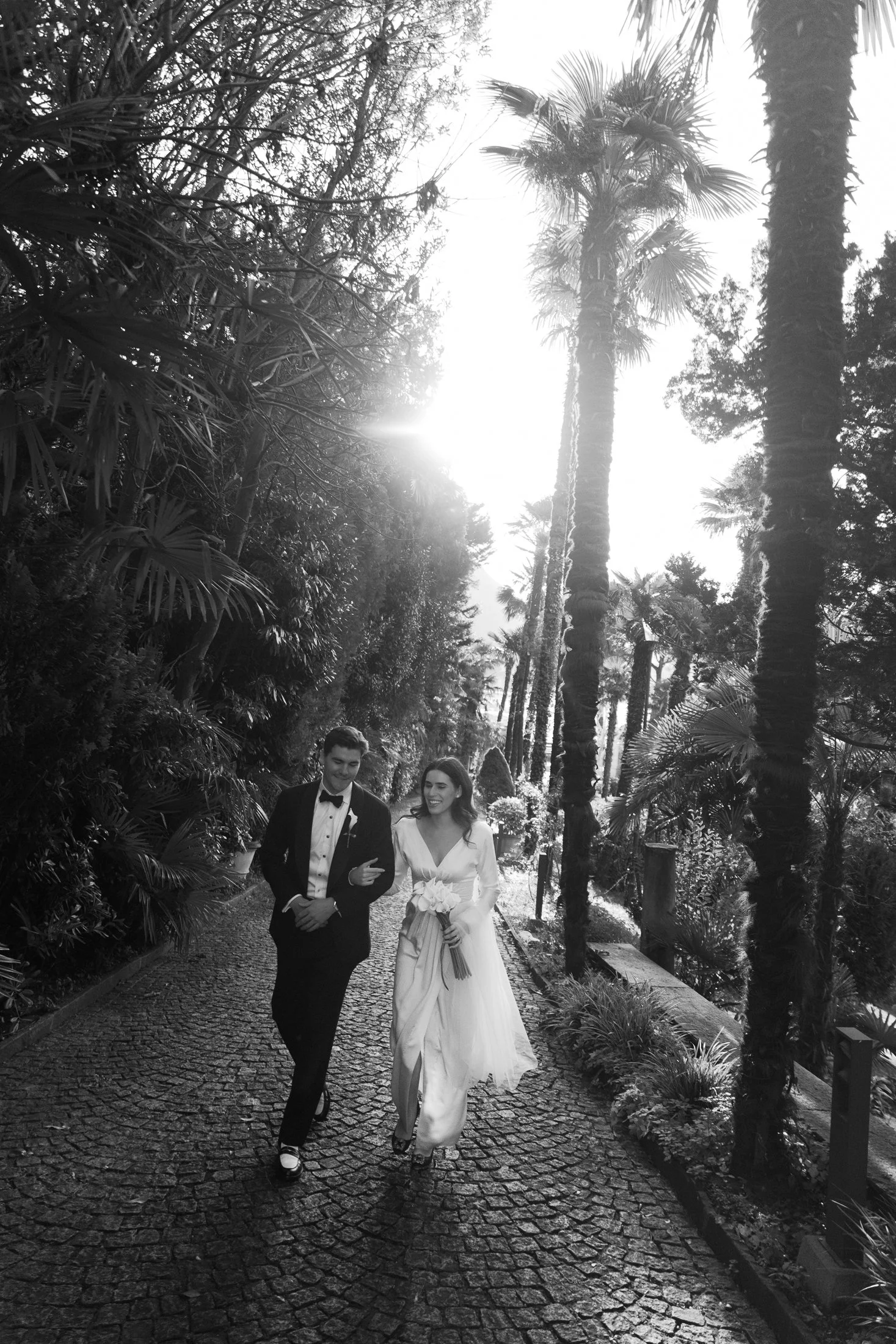 bride and groom happily walking on a path surrounded by palm trees in Lugano