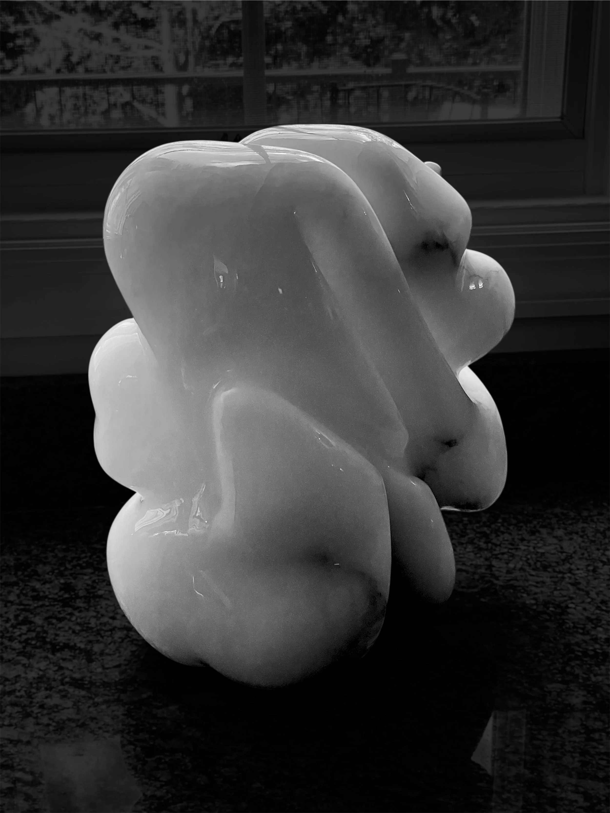 A shiny, abstract, smooth sculpture resembling overlapping rounded shapes, placed on a dark surface near a window with blinds.