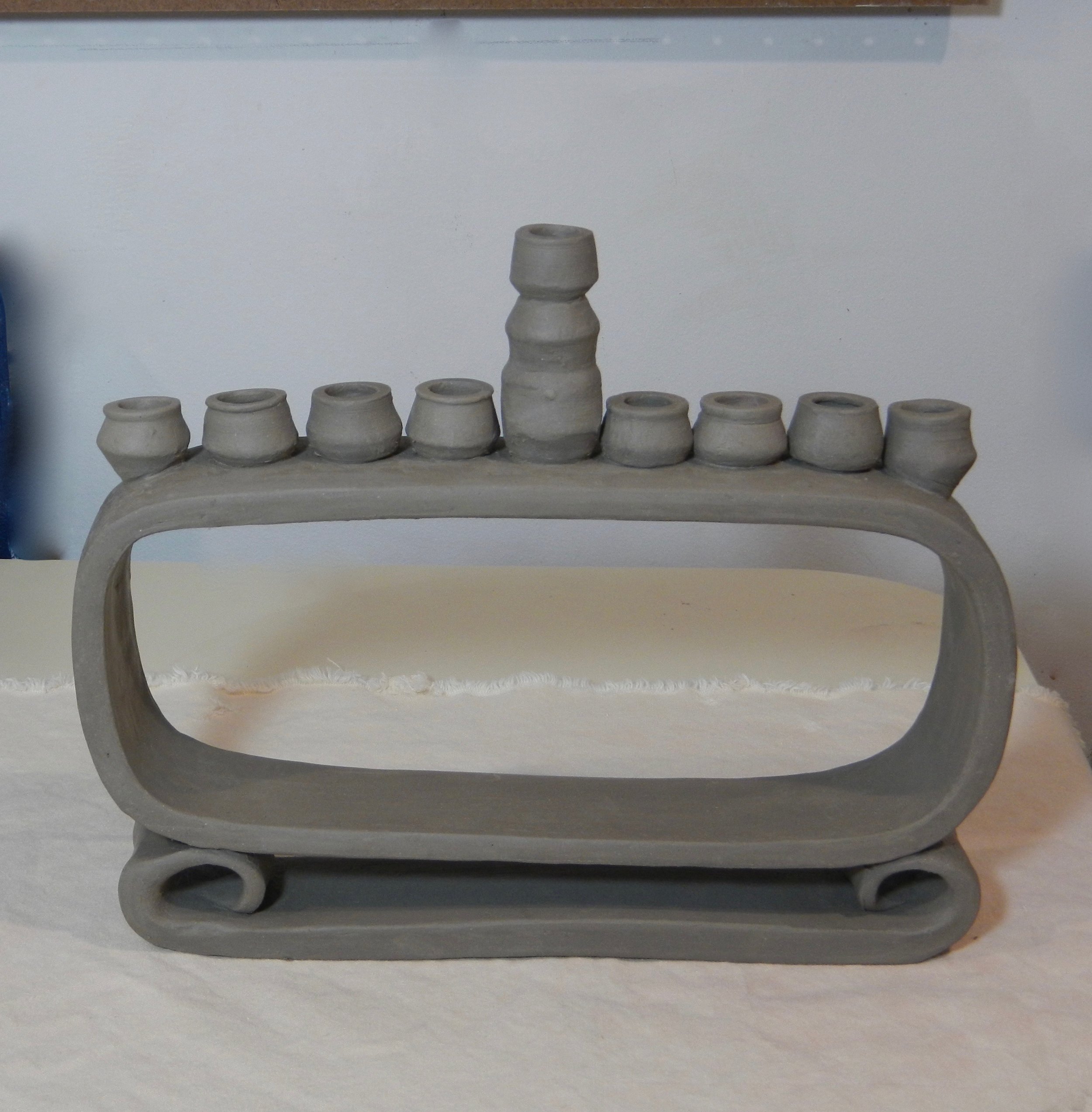 The change here is in the base, a hand-rolled and curled slab with decent thickness instead of a thinner wheel-thrown slab like in #1. The rounded rectangle part is taller. The shamash (the flame that lights all the other lights) is in the center instead of off to the side. (Both shamash placement options are good.) This one is fresh clay.