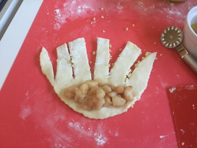 Für die Apfelkringel steche ich ein Rondell aus, bestreiche es mit Butter und streue gehackte Mandeln darüber. Ich drehe das Rondell um und schneide Streifen bis zum oberen Drittel. Ein EL der gekochten Äpfel kommt darauf, dann wickle ich von oben nach unten auf und bilde einen Kreis. 