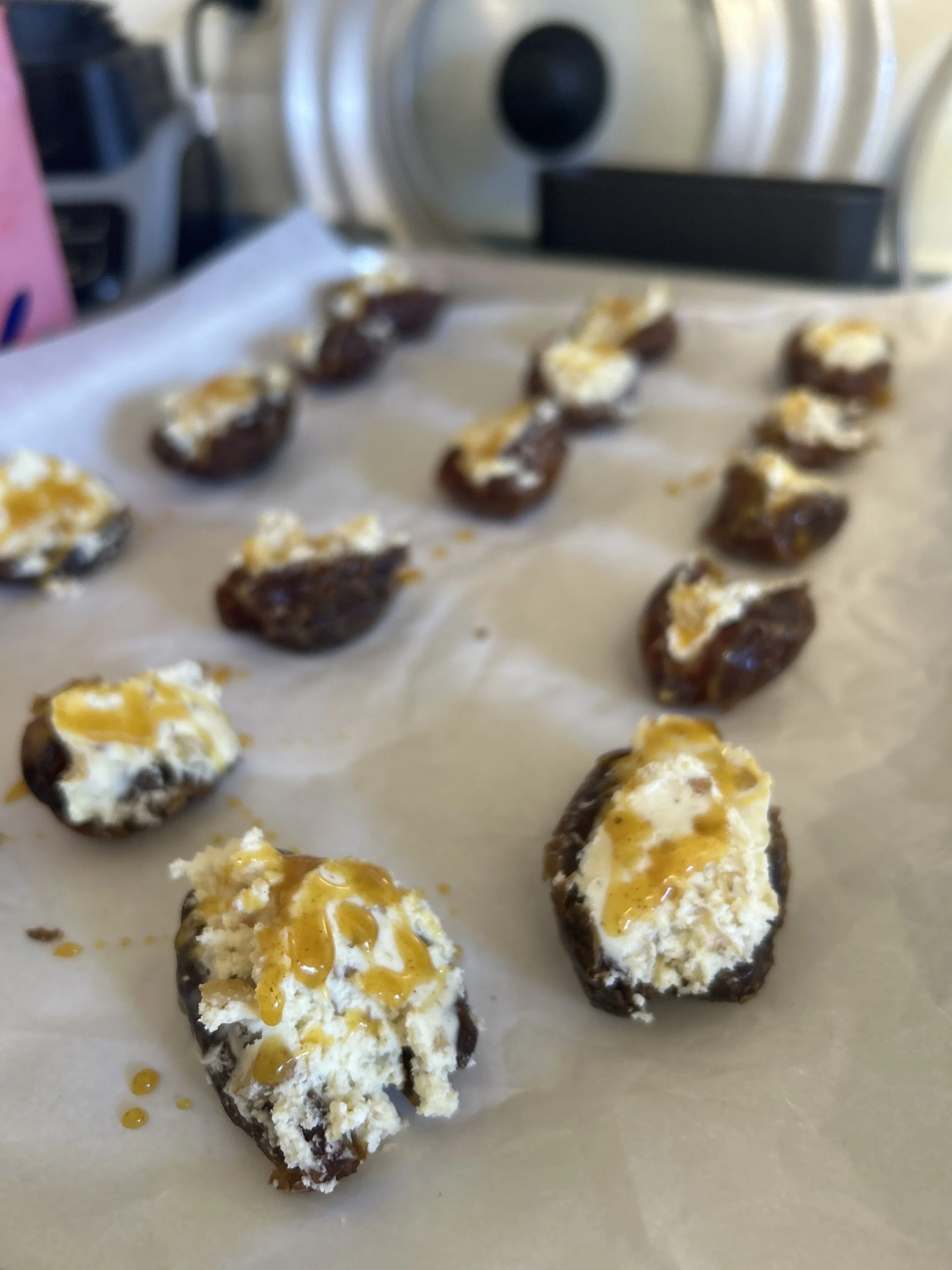 Close-up shot of stuffed dates with goat cheese and honey with blurry ones lined up behind on a stovetop with a large pot lit in the background.