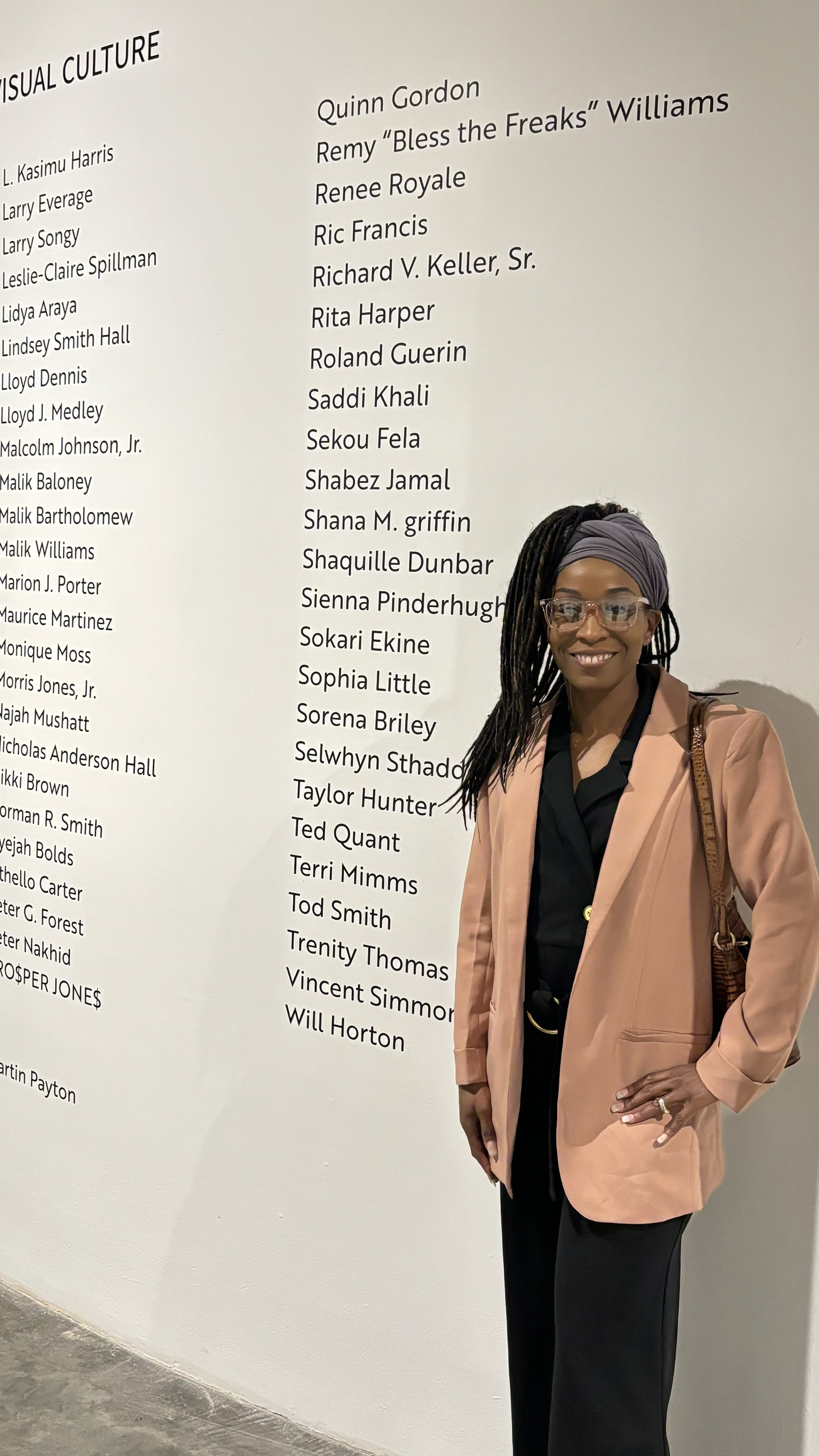 Photographer Sorena Briley smiling in front of the visual culture wall at the Contemporary Arts Center, with her name featured among esteemed artists.