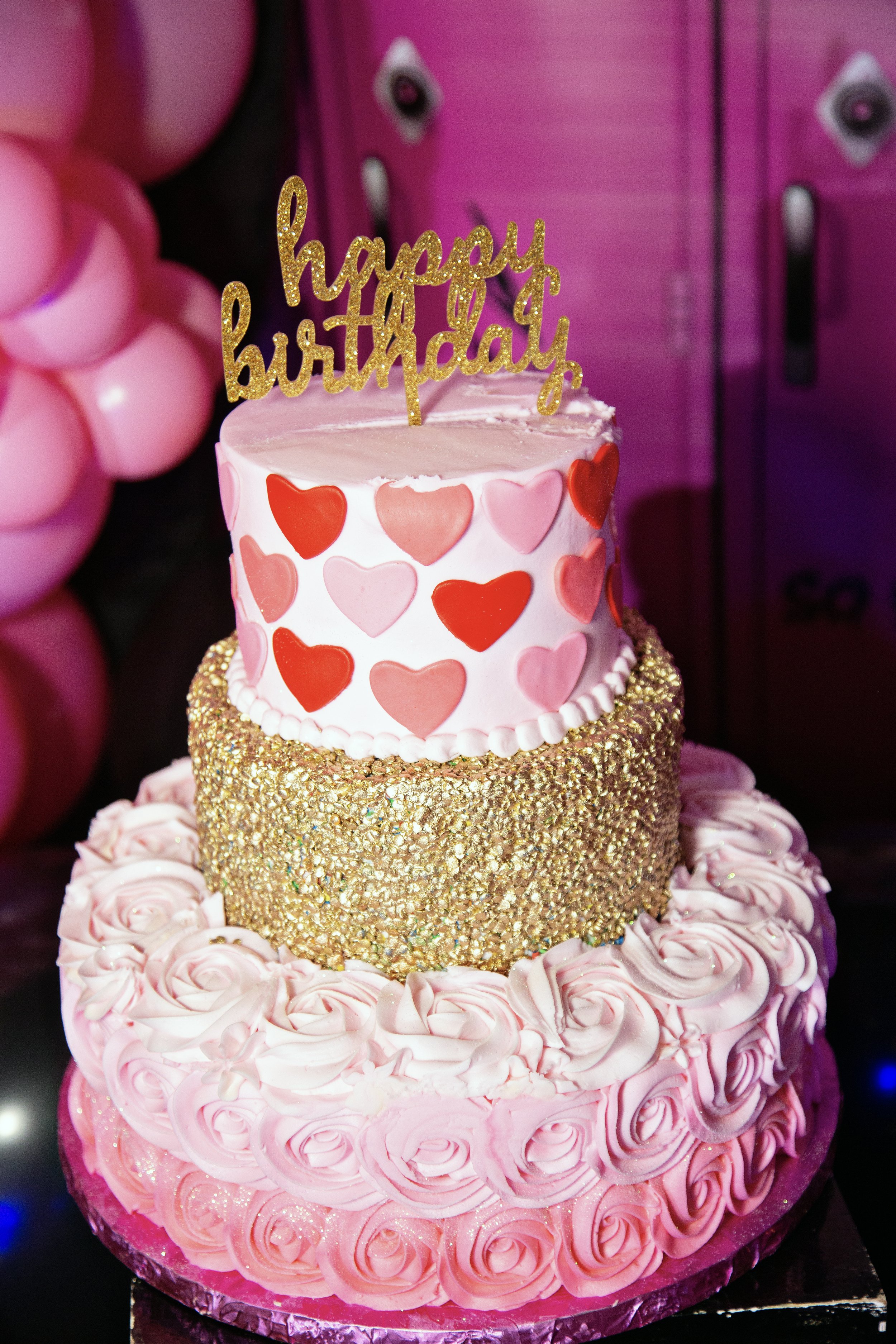 A multi-tiered birthday cake with heart designs and a sparkling golden layer, topped with a 'happy birthday' topper.