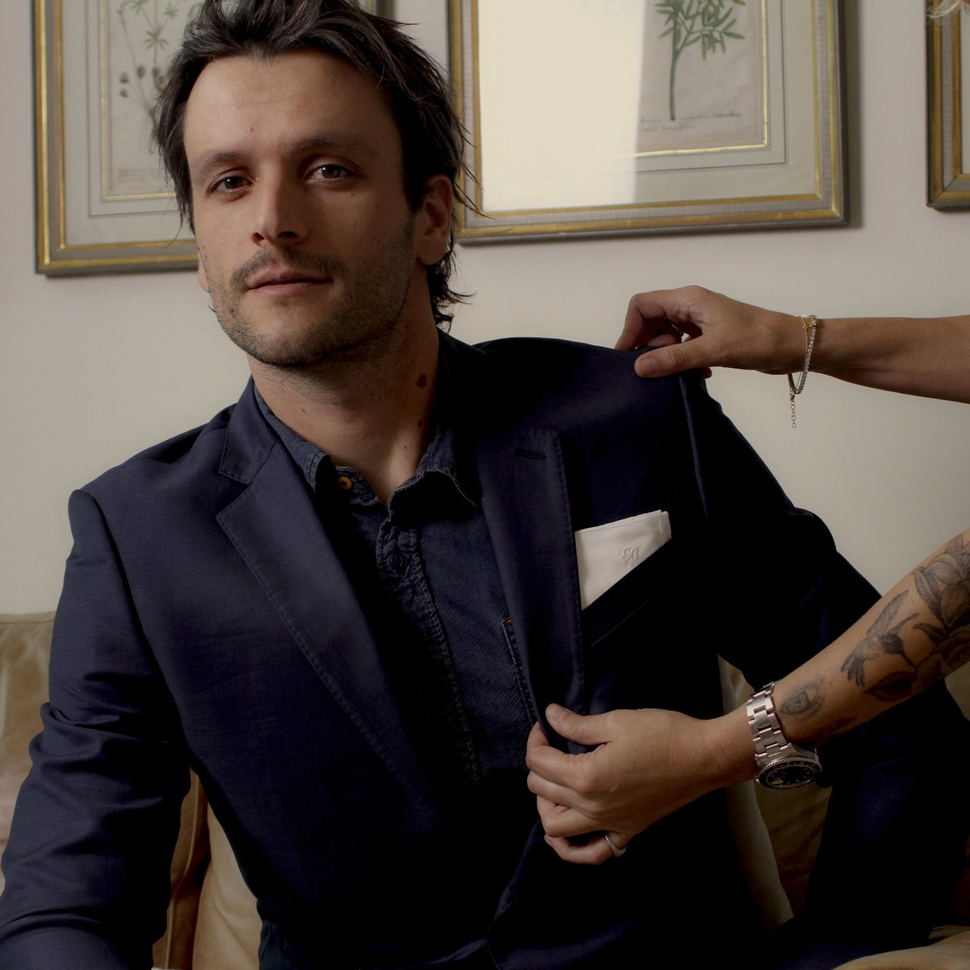Man in a dark blazer being fitted by a tattooed stylist wearing bracelets and a watch