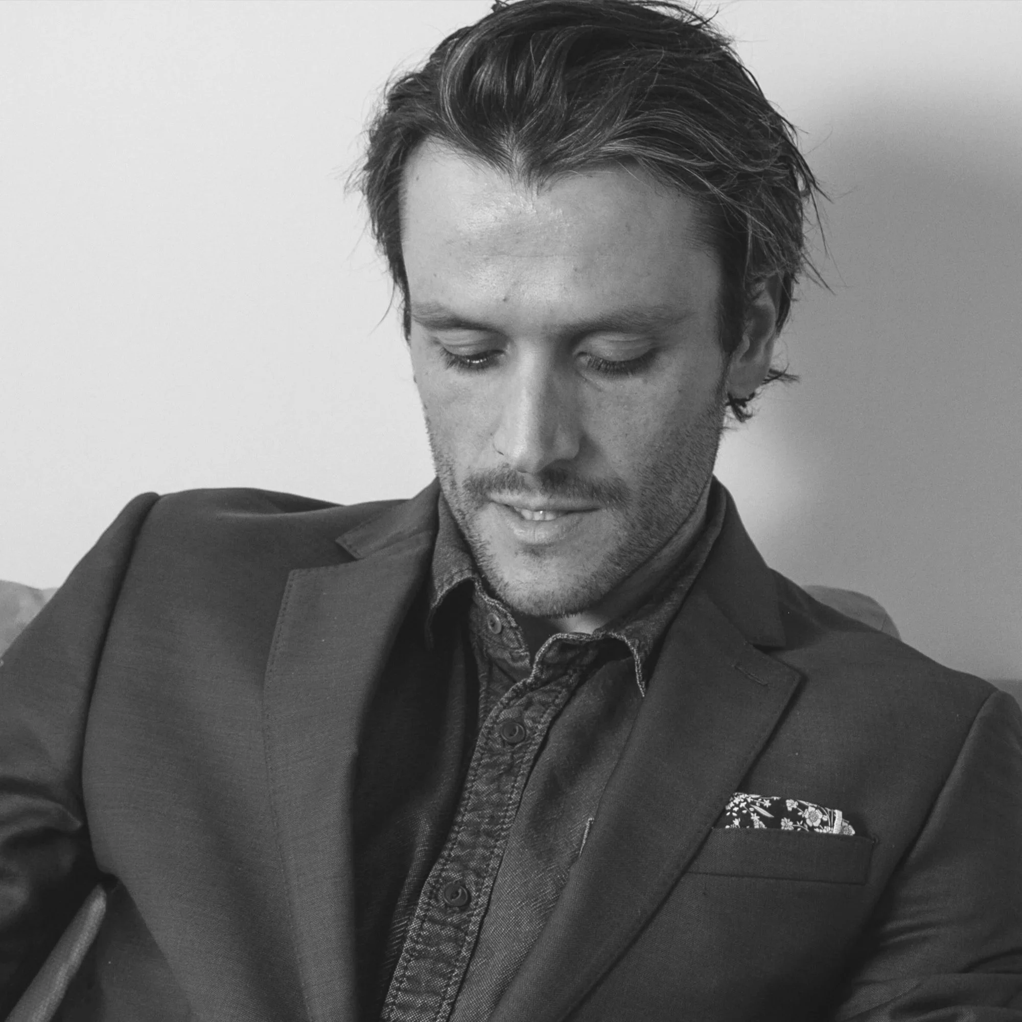 Black-and-white portrait of a bearded man looking down while wearing a blazer, button-up shirt, and patterned pocket square.