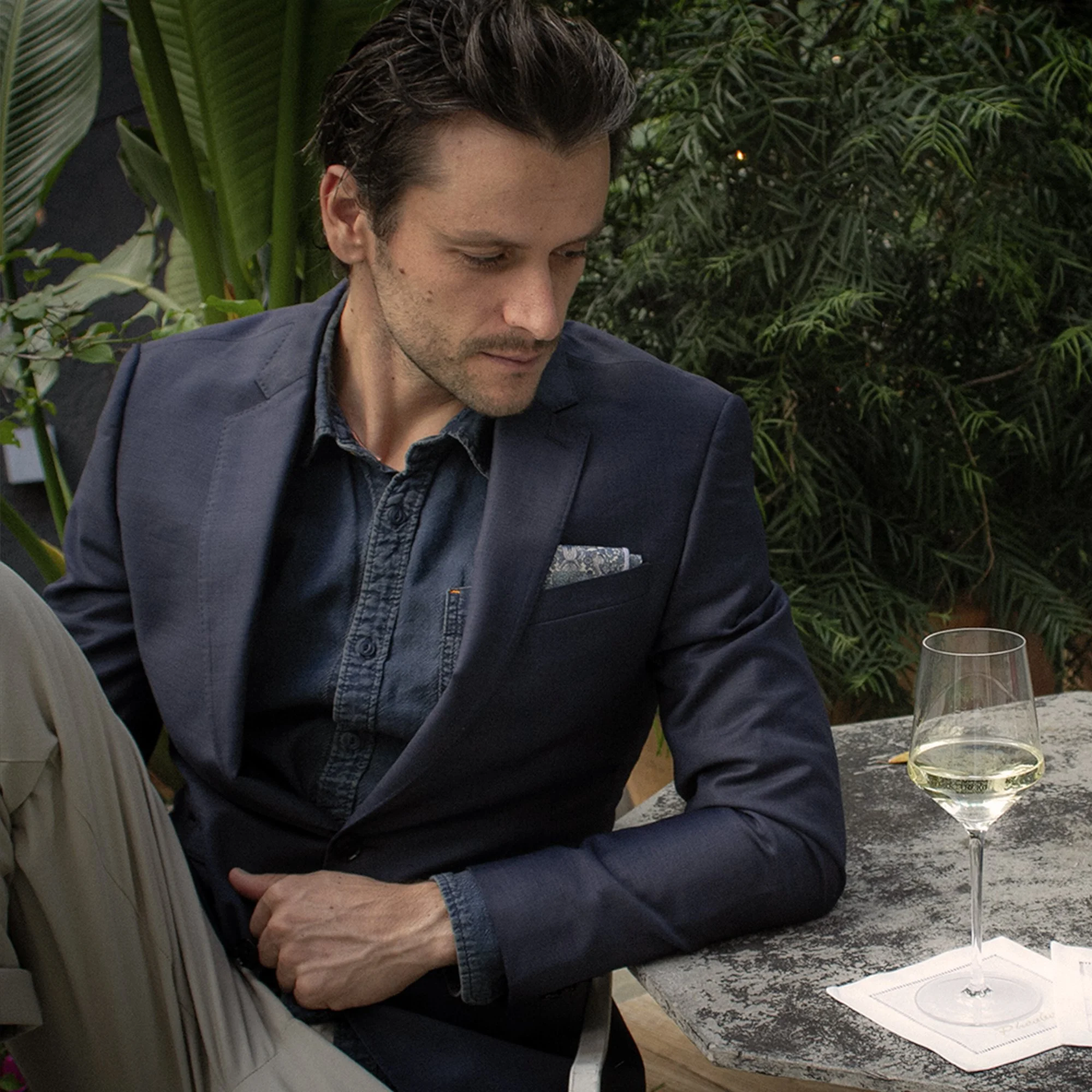 Man in a navy blazer seated at an outdoor restaurant table with a glass of white wine and lush plants behind him