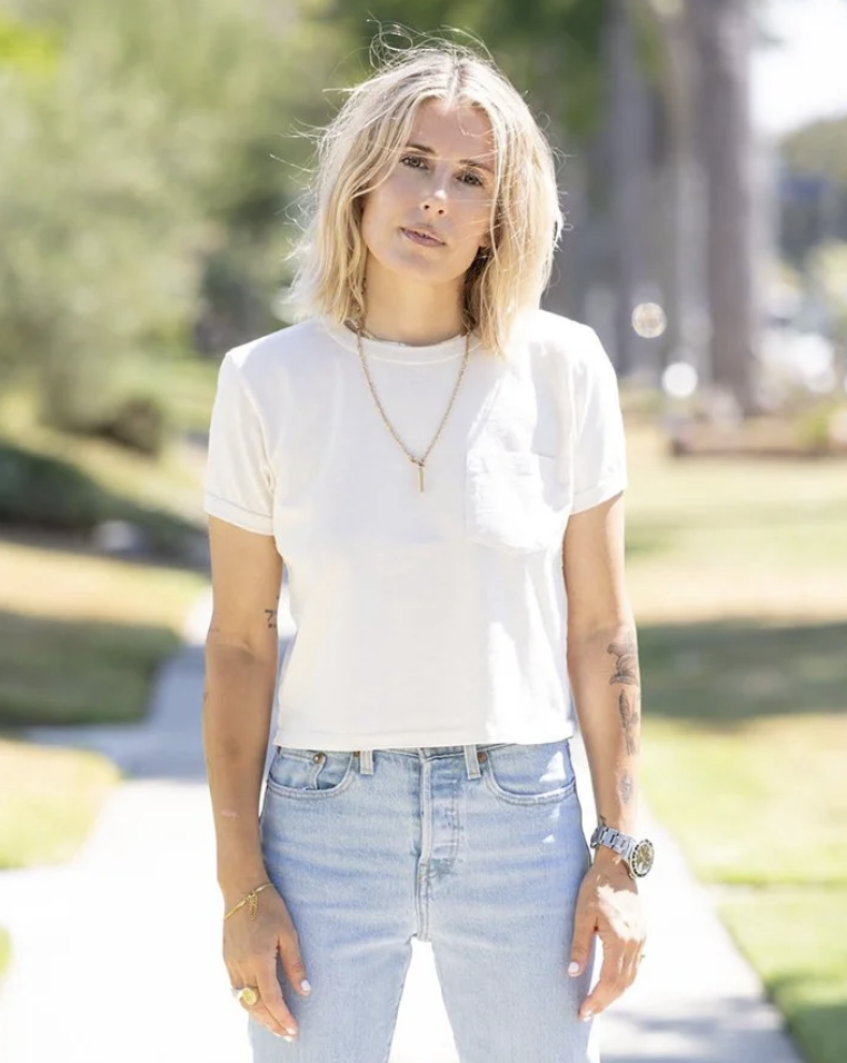 A young woman with shoulder-length blonde hair standing outdoors on a sunny day, dressed in a white t-shirt and light blue jeans.