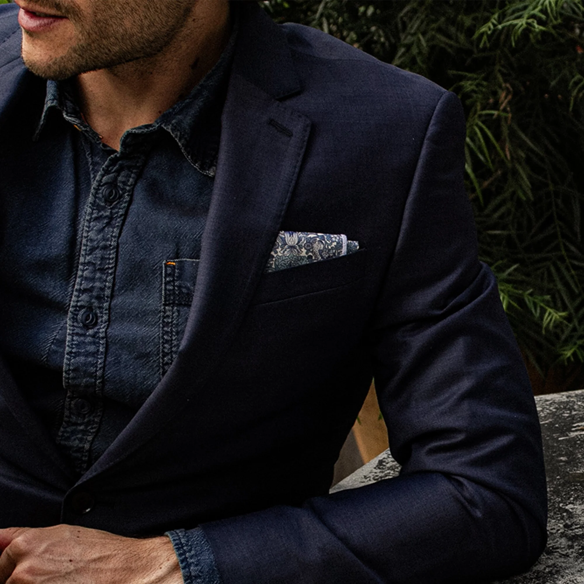 Close-up of a man wearing a navy blazer, dark denim shirt, and patterned pocket square outdoors