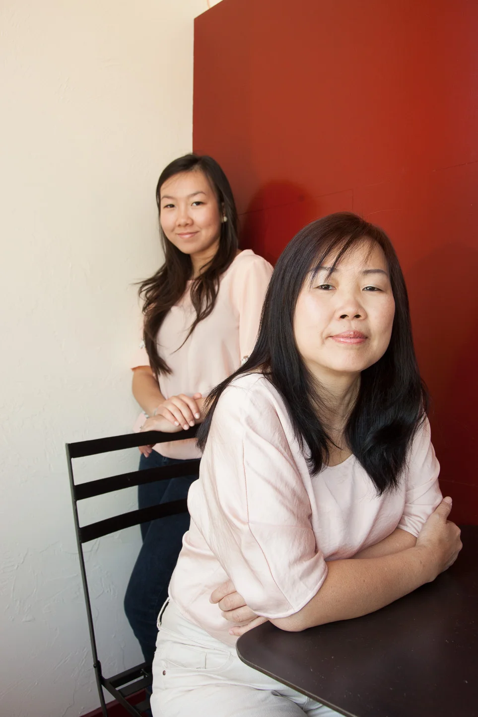 Two women with black hair in light pink tops, one sitting at a black table and the other standing behind her, against a white wall and red panel background.