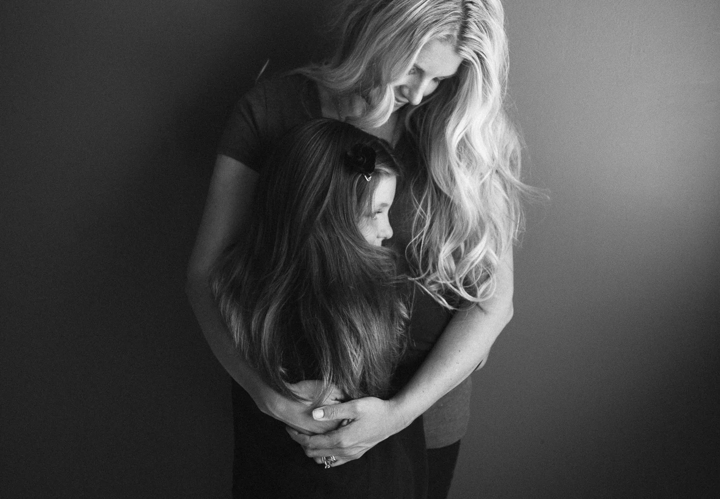 A woman with long, wavy blonde hair embraces a young girl with long, dark hair, both looking down, in a black and white photo.