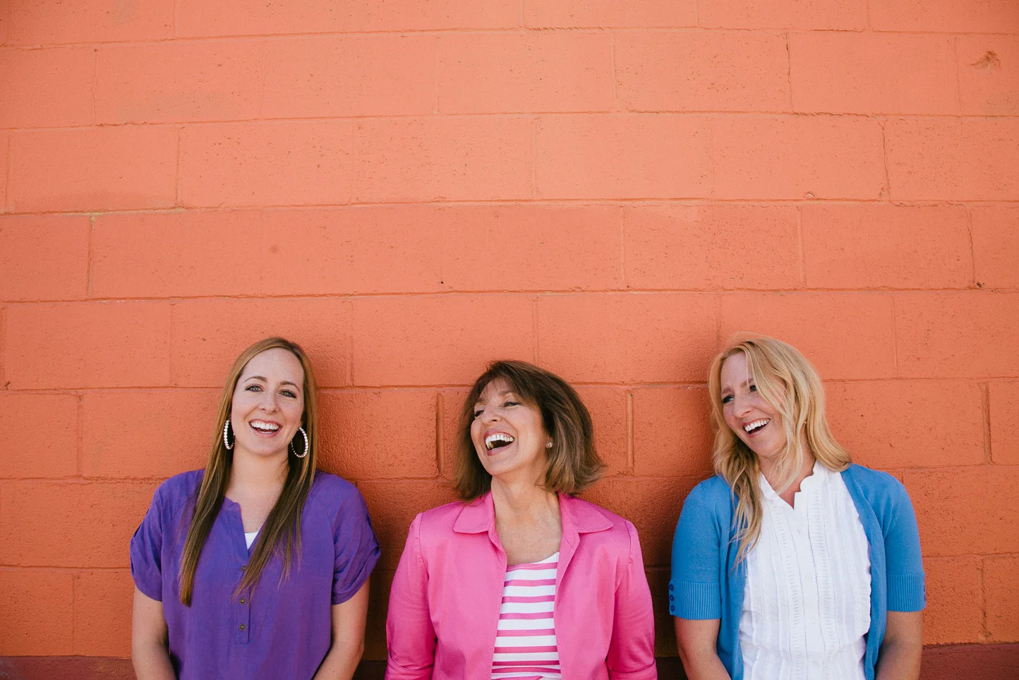 Three women standing in front of an orange brick wall, smiling and laughing. The woman on the left wears a purple blouse, the woman in the middle wears a pink jacket with a striped shirt underneath, and the woman on the right wears a blue cardigan ov