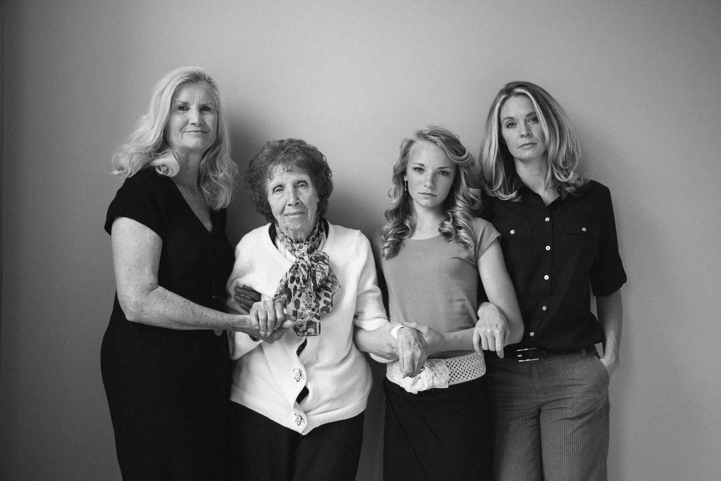 Black and white photo of four women standing against a plain wall, with the oldest woman in the center holding a cane and wearing a scarf. The women vary in age, from elderly to young adult, with serious expressions.