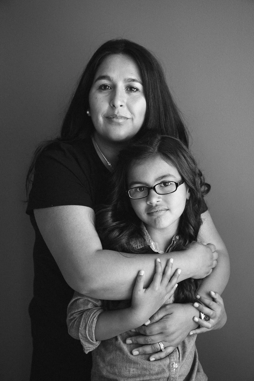 A black and white photo of a woman with her arms around a young girl with glasses, both looking at the camera.