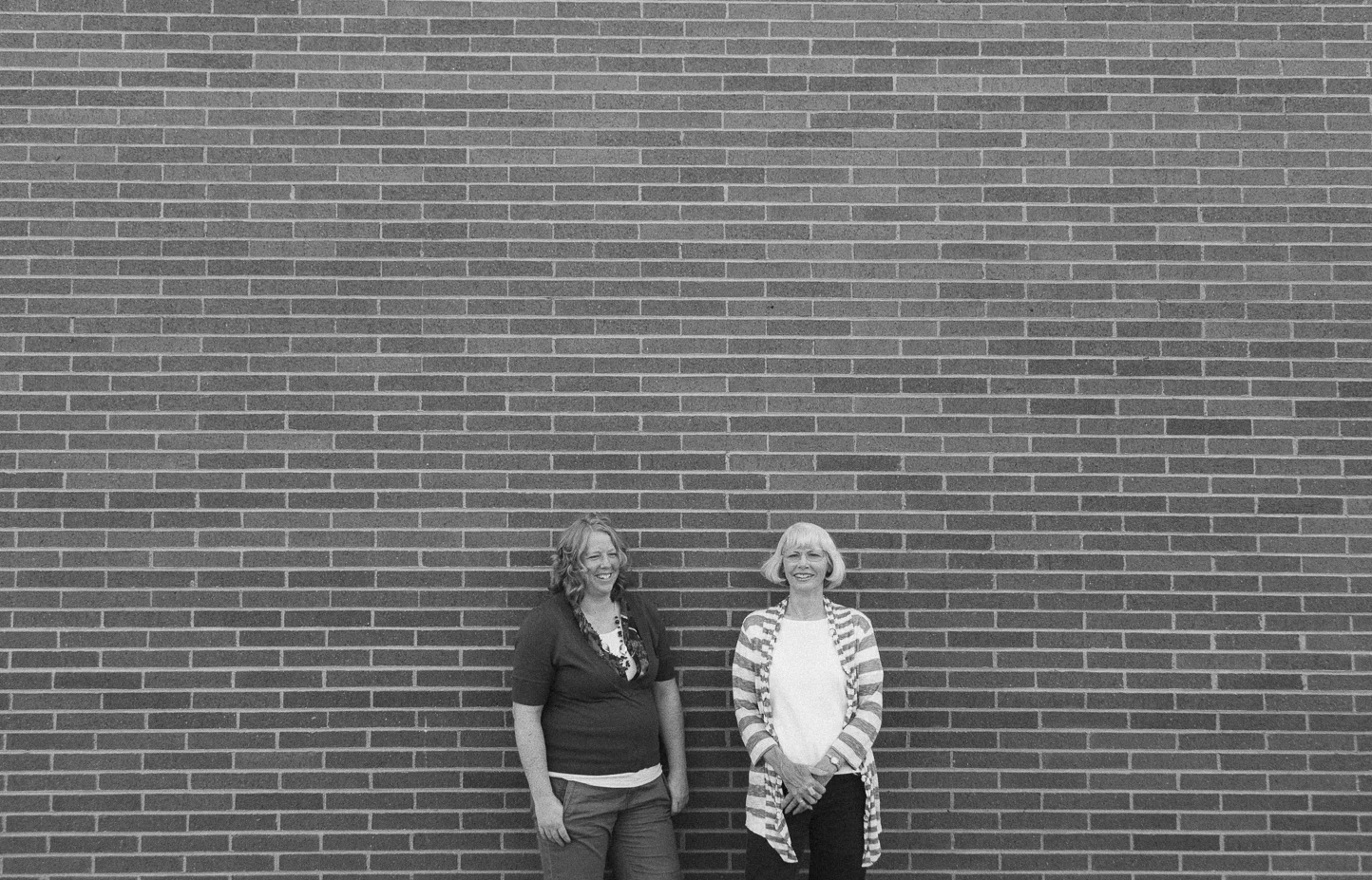 Two women standing against a large brick wall, smiling and looking at each other.