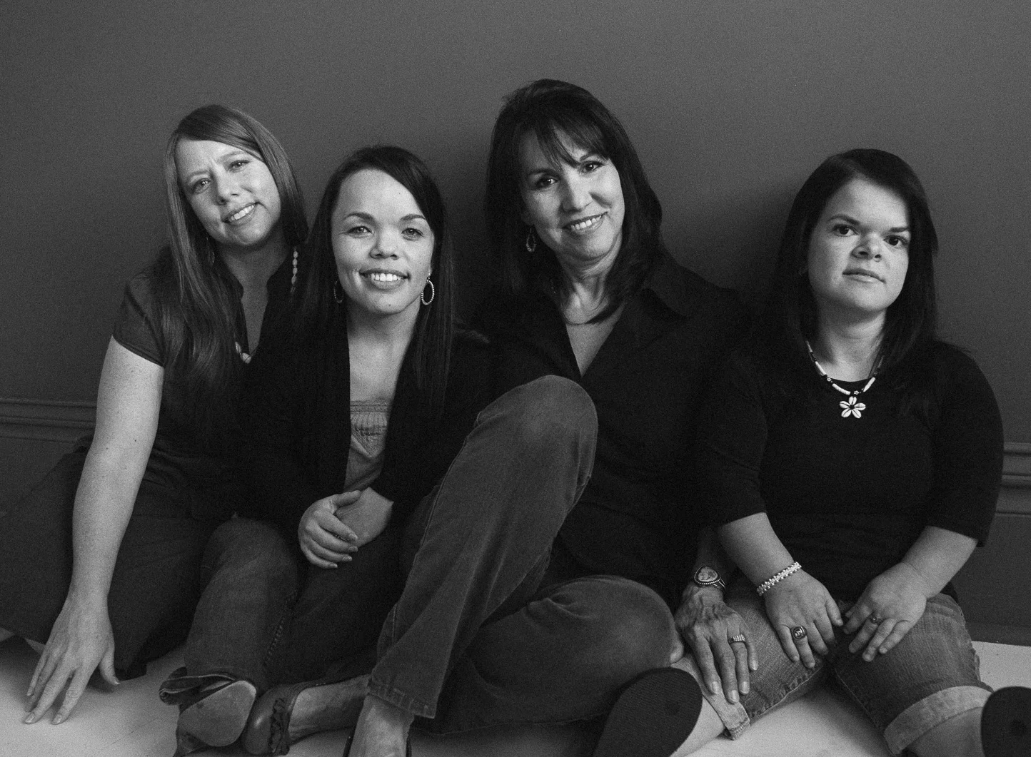 Black and white photo of four women sitting close together against a plain wall, smiling at the camera.