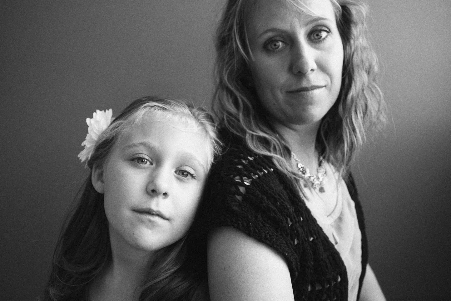 Black and white portrait of a young girl with long hair and a flower in her hair, leaning her head on an older woman's shoulder. The older woman has wavy hair and is wearing a necklace and a black, open-knit top.