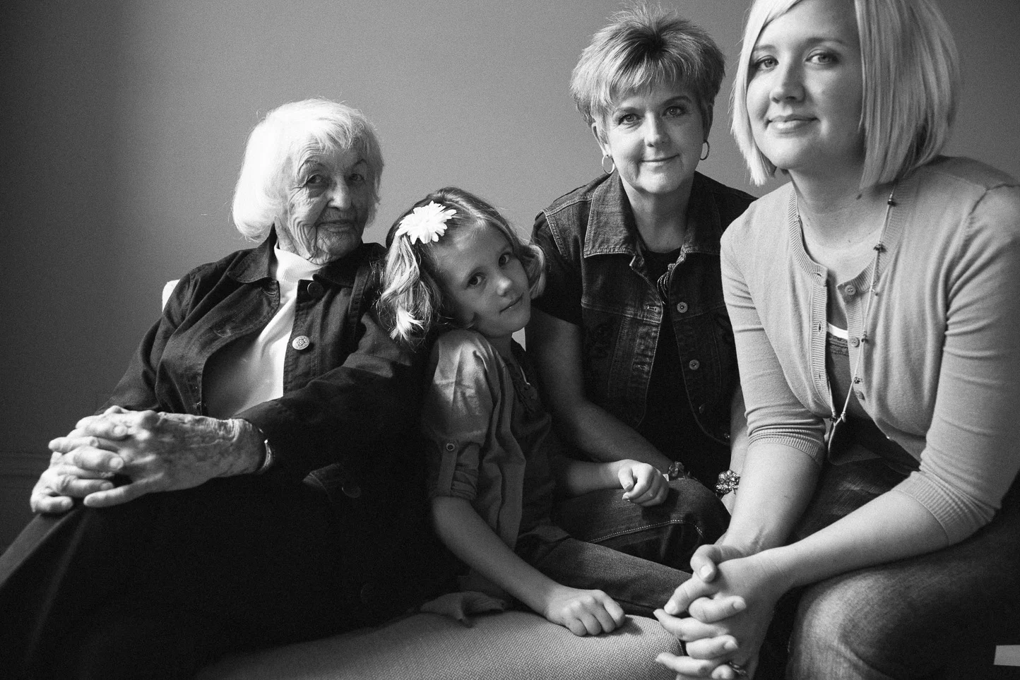 Black and white photo of four generations of women sitting together on a couch: an elderly woman, two middle-aged women, and a young girl, all smiling at the camera.