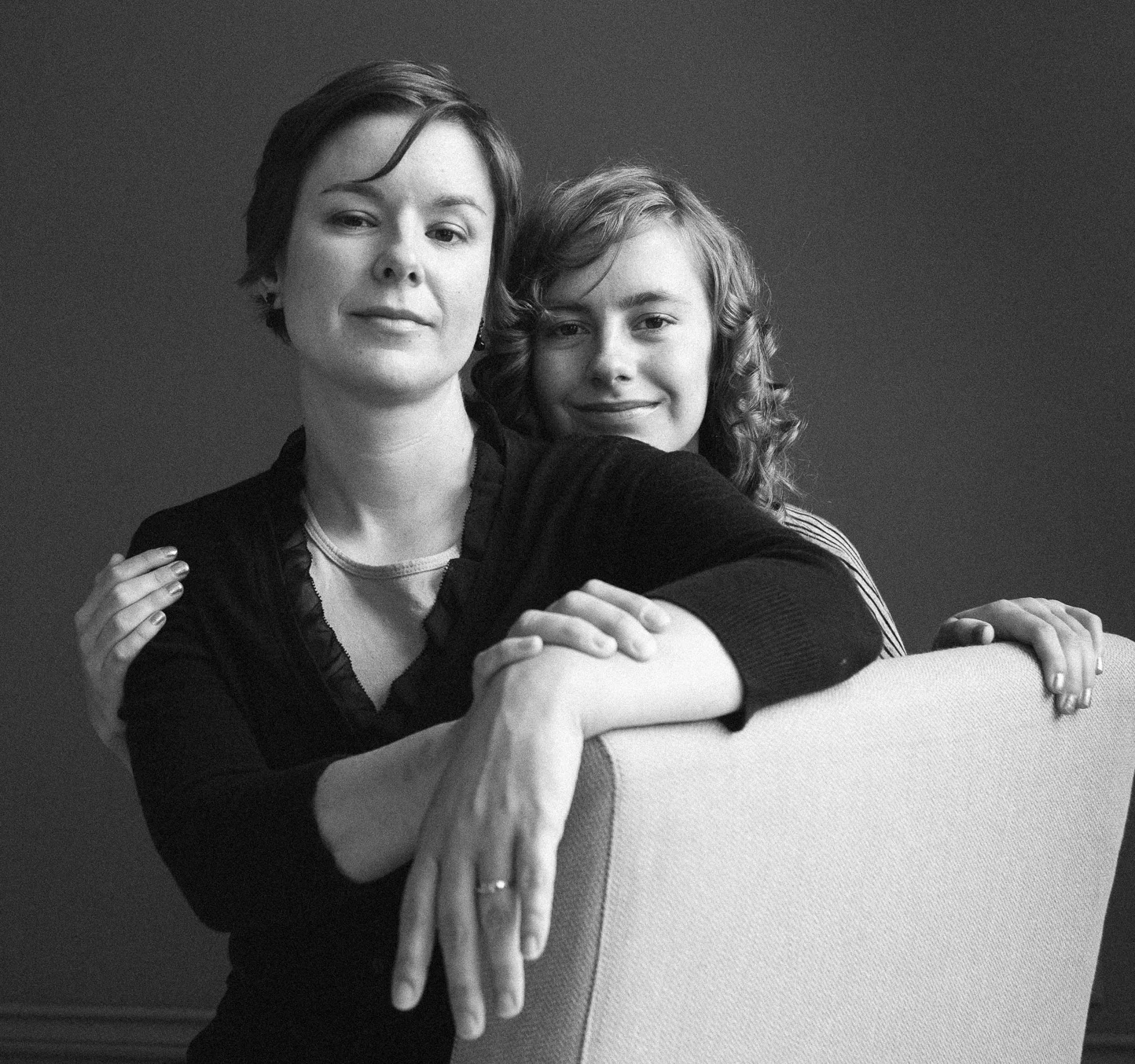 A black and white portrait of two women, one with short dark hair and the other with curly hair, sitting close together with one resting her arms on the back of a chair, smiling gently at the camera.