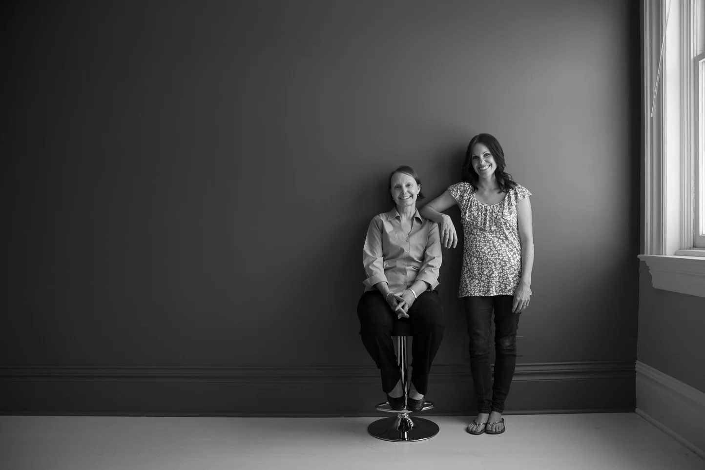 Black and white photo of two women in a room with plain wall and window, one sitting on a stool and the other standing beside her with arm resting on the seated woman's shoulder, both smiling.