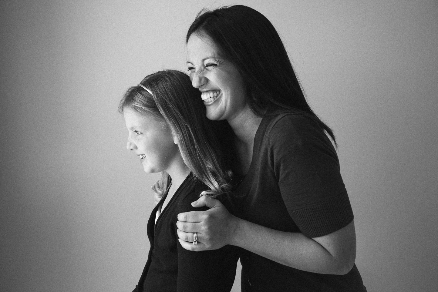 A woman and a young girl smiling and enjoying a moment together, the woman embracing the girl from behind.