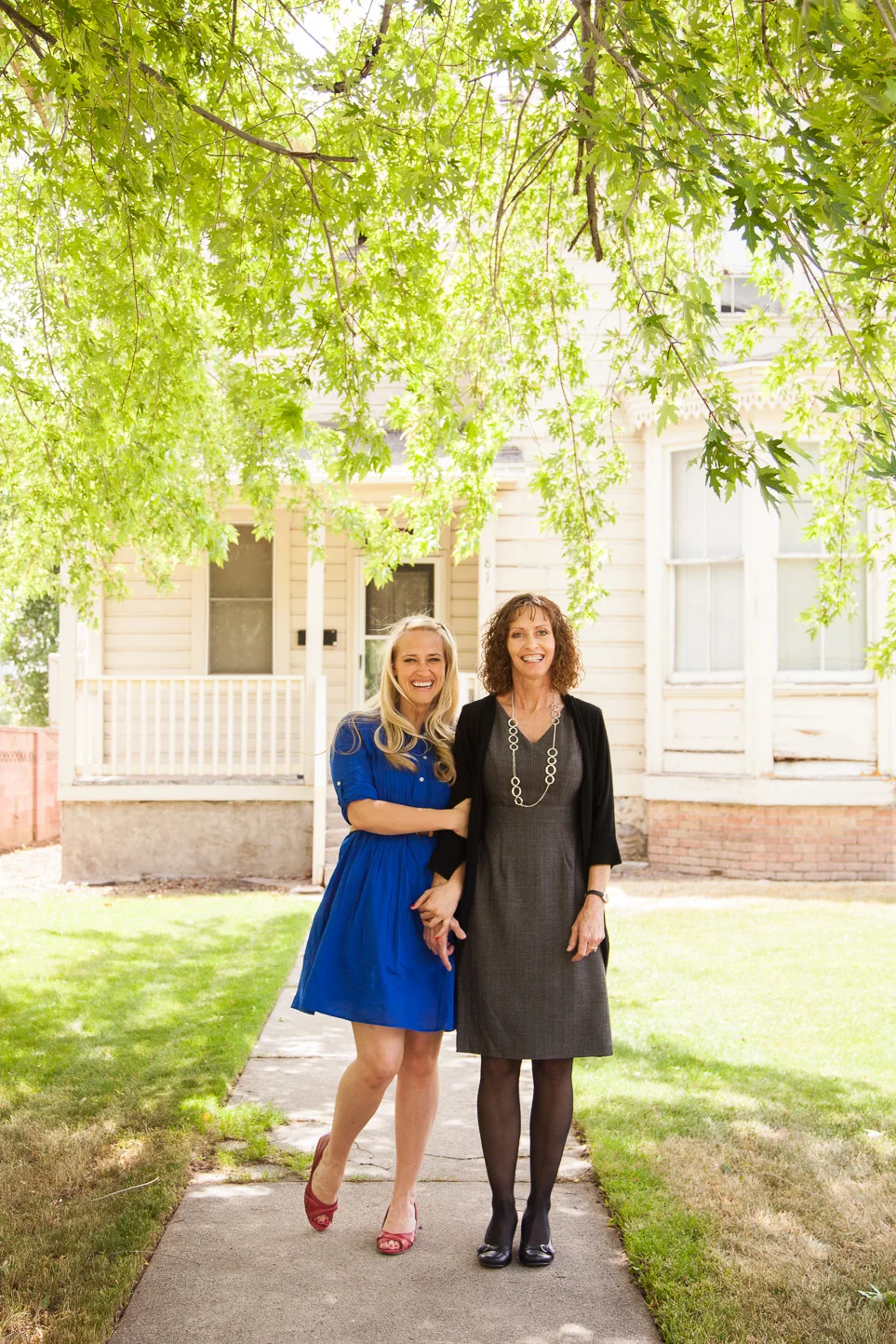 Two women standing on a sidewalk in front of a house, holding hands and smiling. The woman on the left has long blonde hair, is wearing a blue dress and red shoes. The woman on the right has curly brown hair, is dressed in a gray dress with black tig
