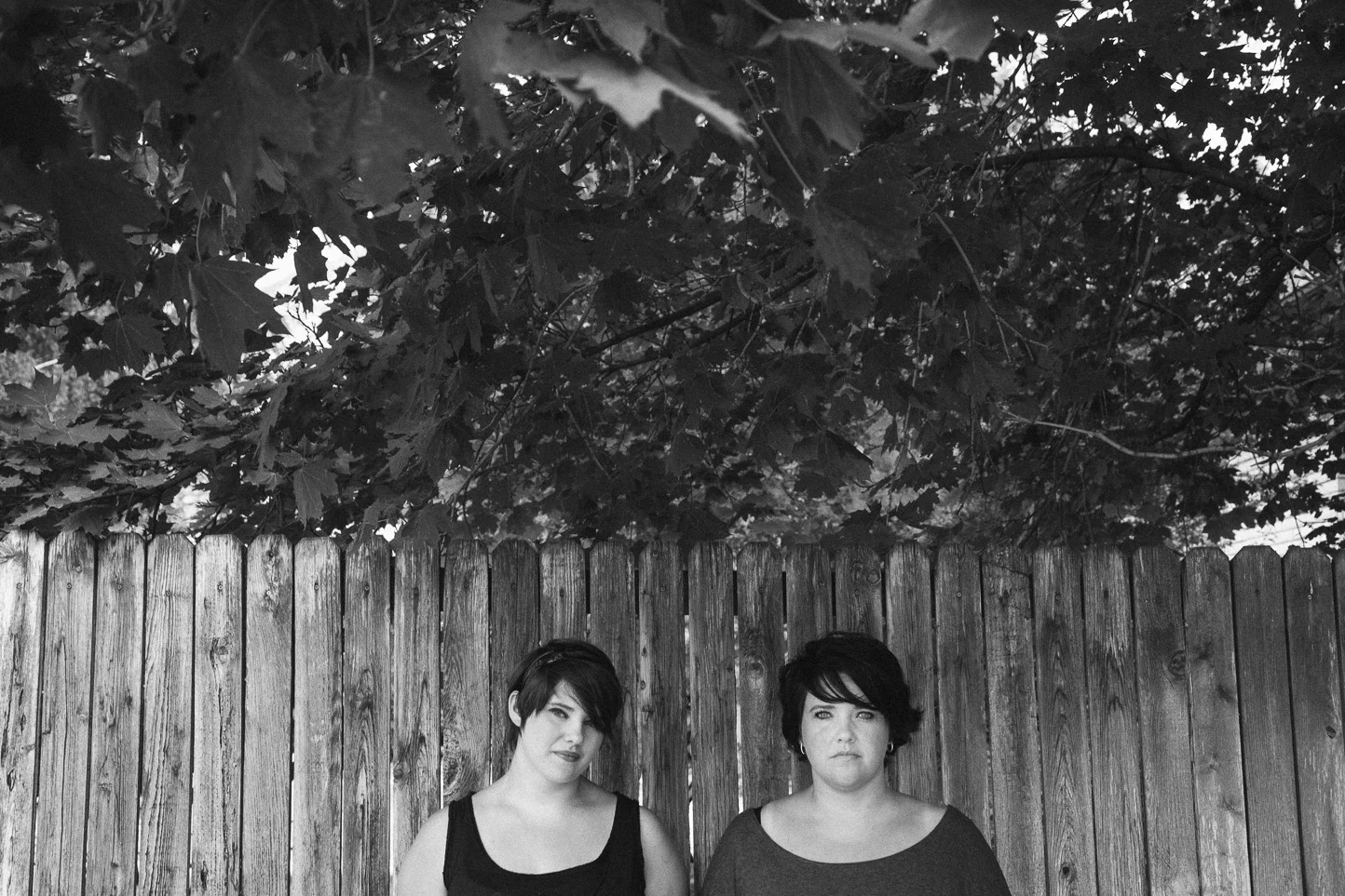 Black and white photo of two women with short dark hair standing in front of a wooden fence beneath overhanging tree branches.