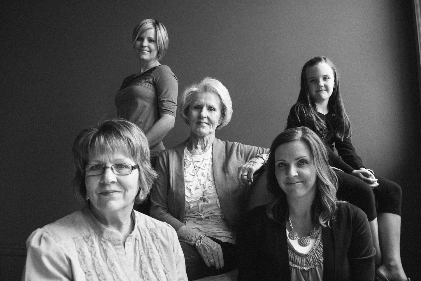 Black and white portrait of six women and girls, posing together against a plain wall, sitting and standing in a close group.