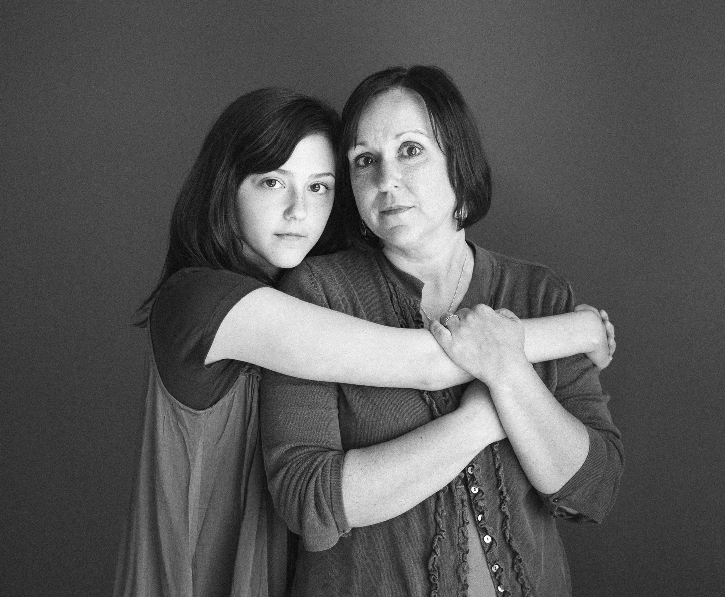 A young girl and an older woman, possibly her mother, embracing each other against a plain background. The girl has long dark hair and the woman has short dark hair, and there is a tender connection between them.