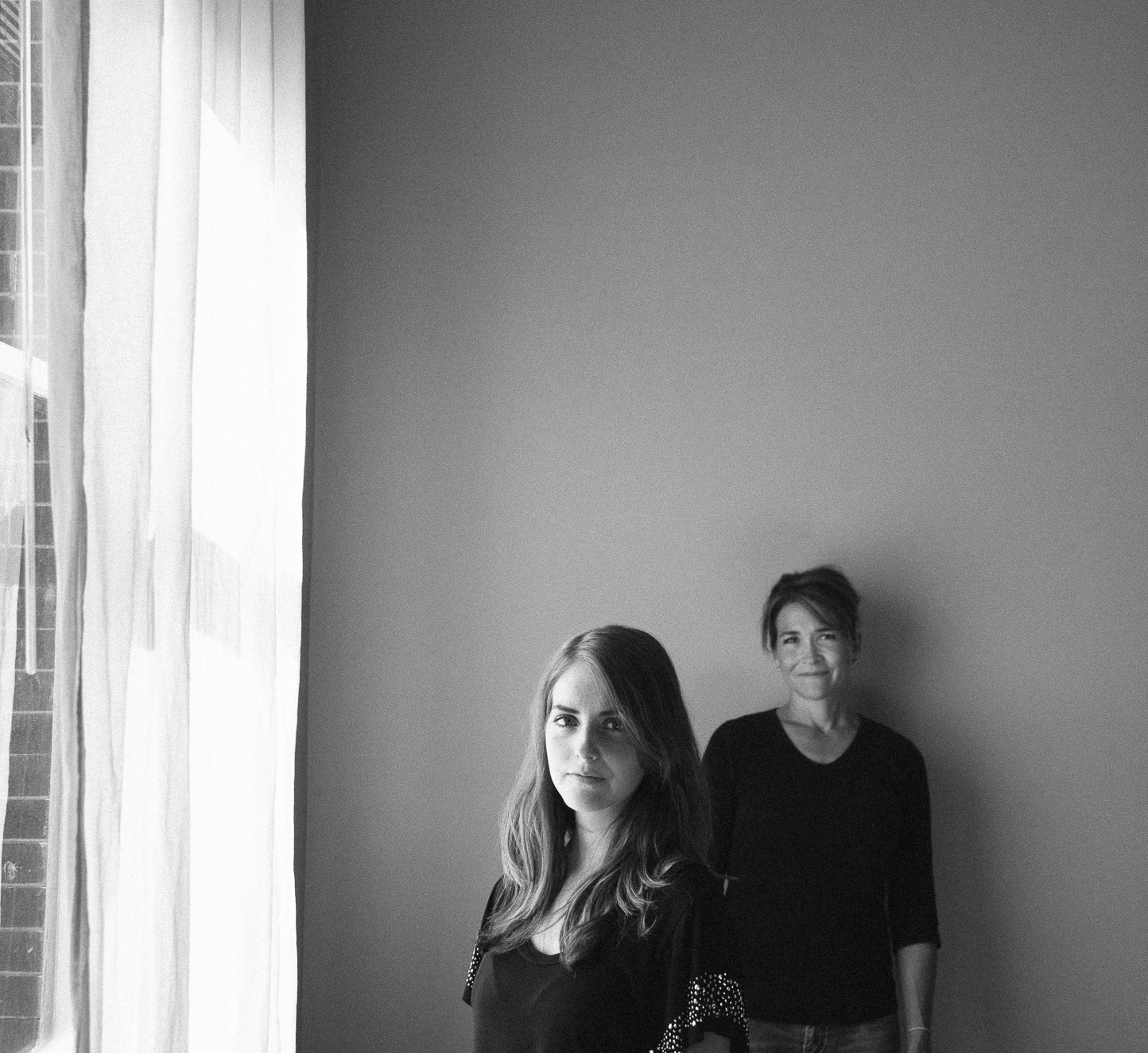 Two women standing in a room near a large window with sheer curtains, one woman with long hair in the foreground and the other with short hair smiling in the background, both dressed in dark clothing.