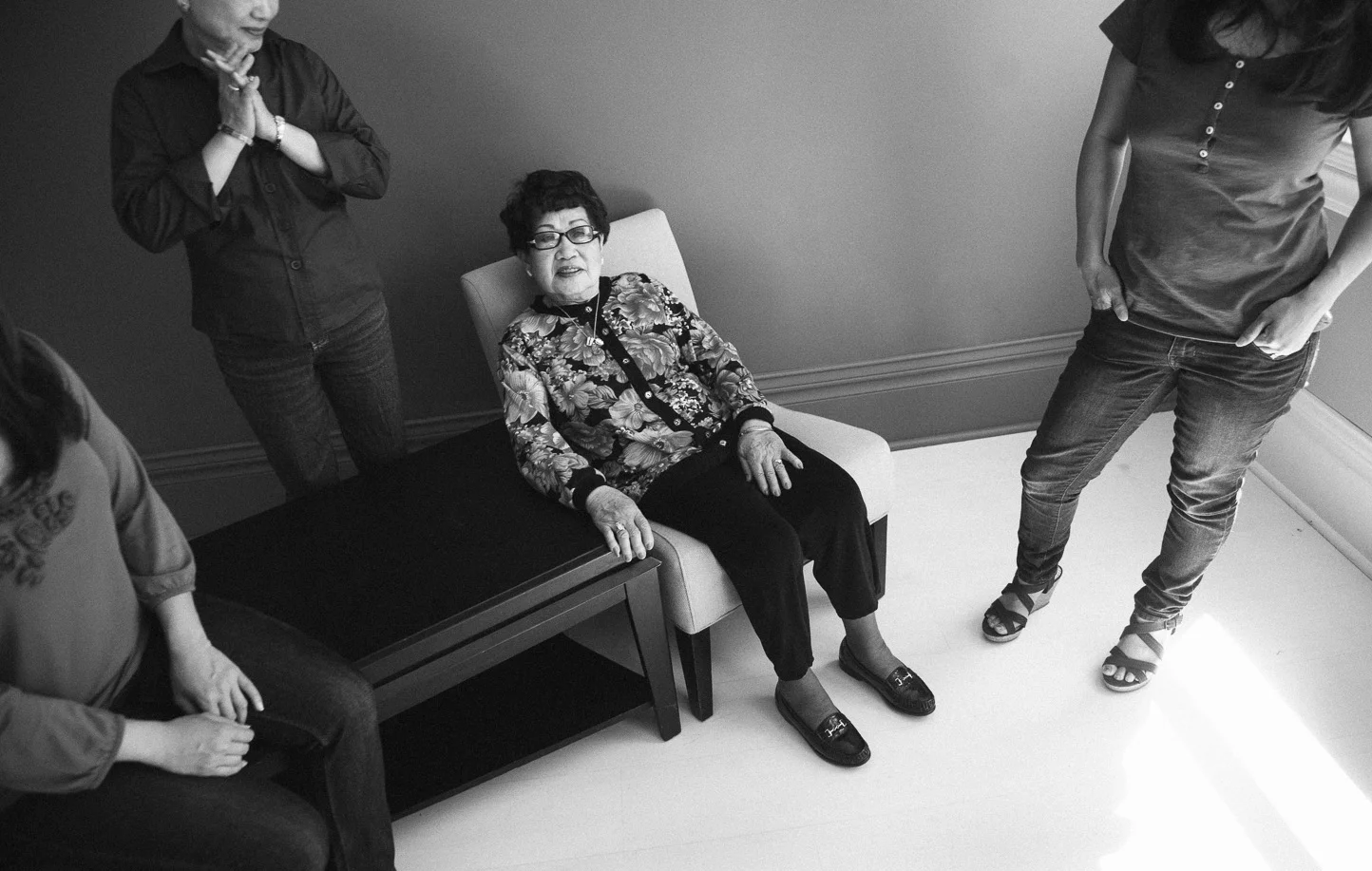 An elderly woman with glasses sitting on a chair surrounded by three younger women in a room with plain walls and light flooring.