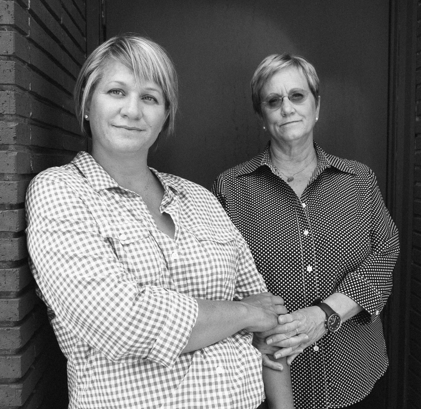 Black and white photo of two women standing against a brick wall. The woman on the left has short hair, wears a checkered shirt, and has her arms crossed. The woman on the right has short hair, wears glasses and a polka dot shirt, and is holding the 