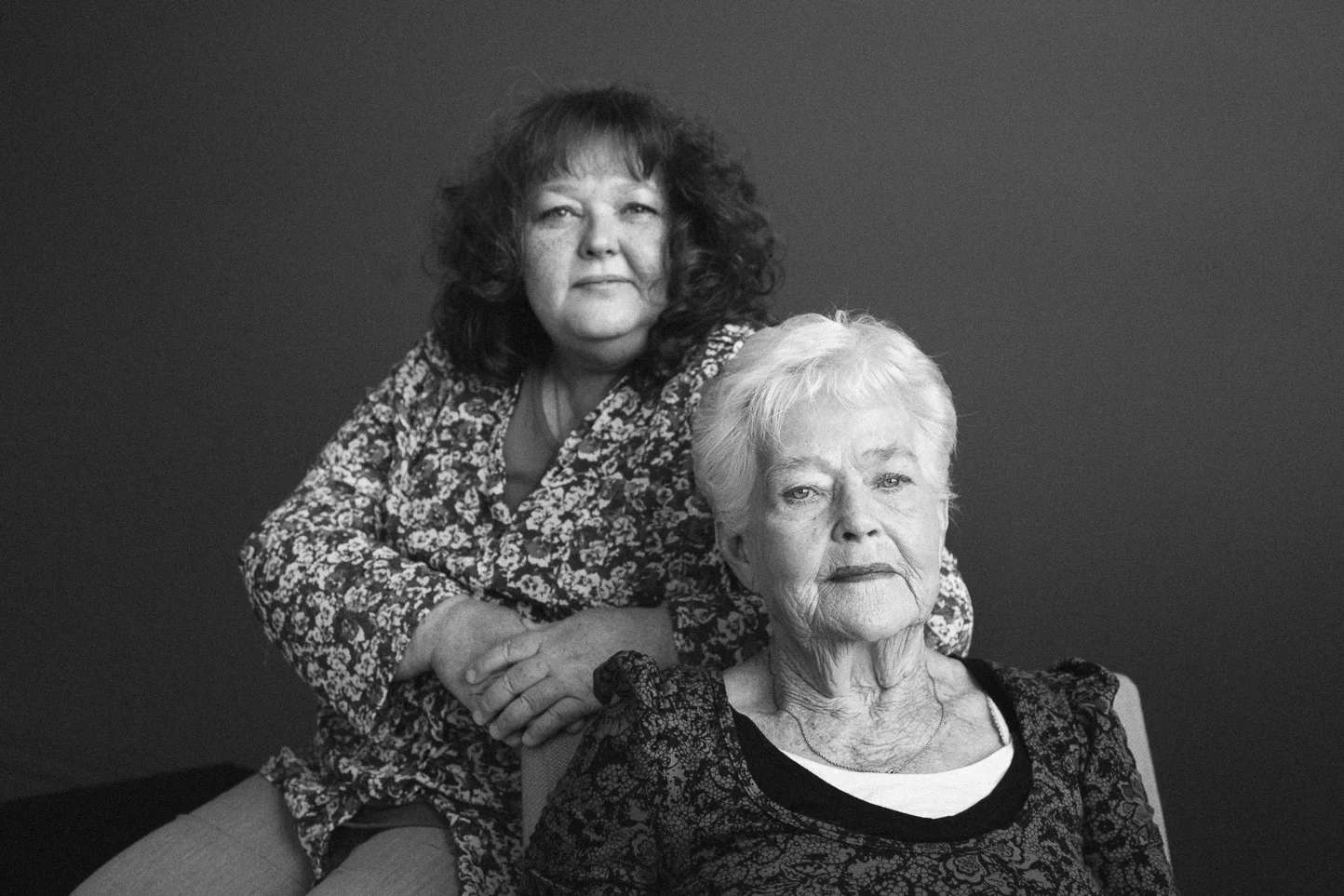 Black and white portrait of two older women, one sitting in front with short white hair and a serious expression, the other standing behind with curly dark hair and a neutral expression, against a plain dark background.
