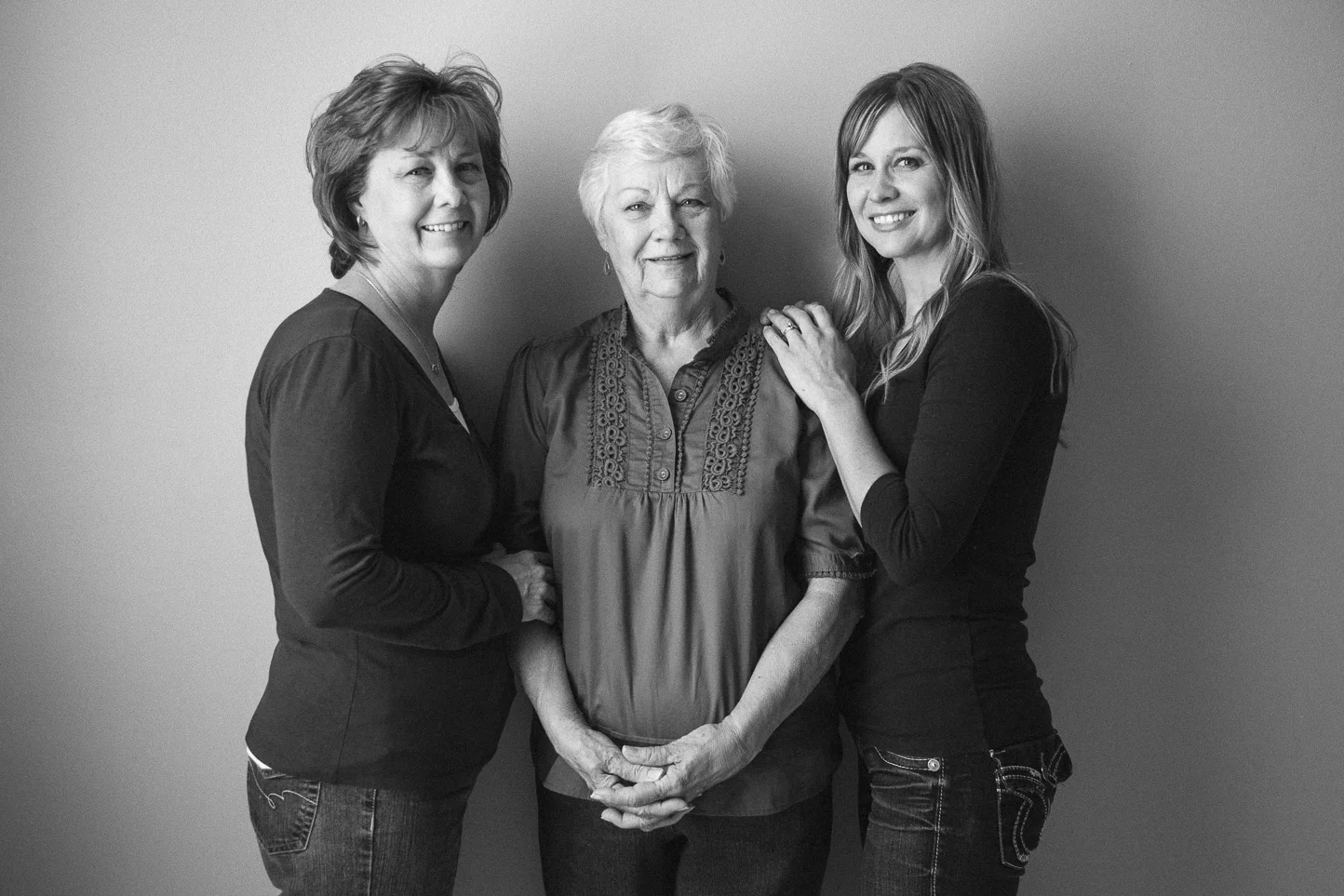 Black and white photo of three women standing close together, smiling at the camera, against a plain wall. The woman in the middle is elderly with short gray hair, wearing a decorative blouse. The woman on the right has long blonde hair and is touchi