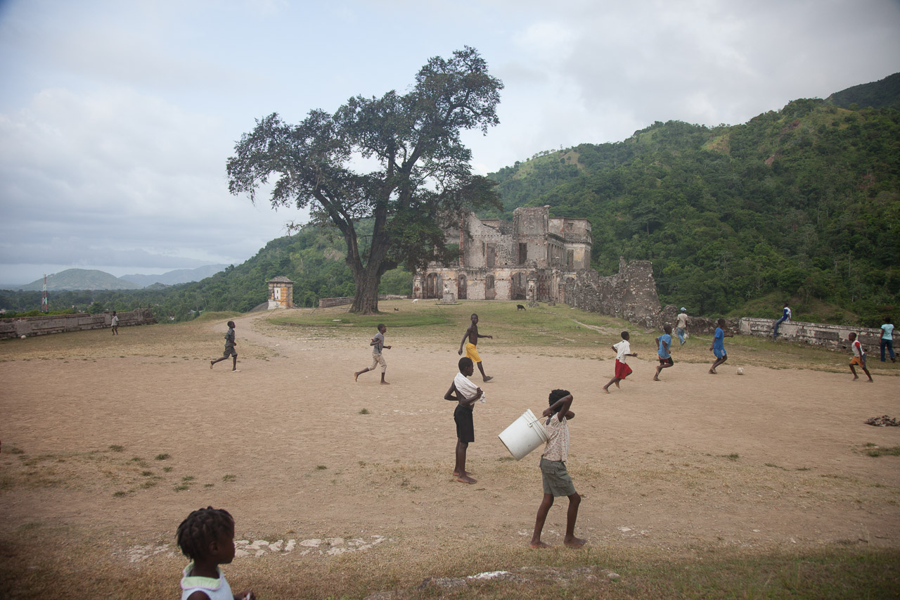Children playing soccer on a dust field with a large tree, old stone building, and green mountain backdrop.