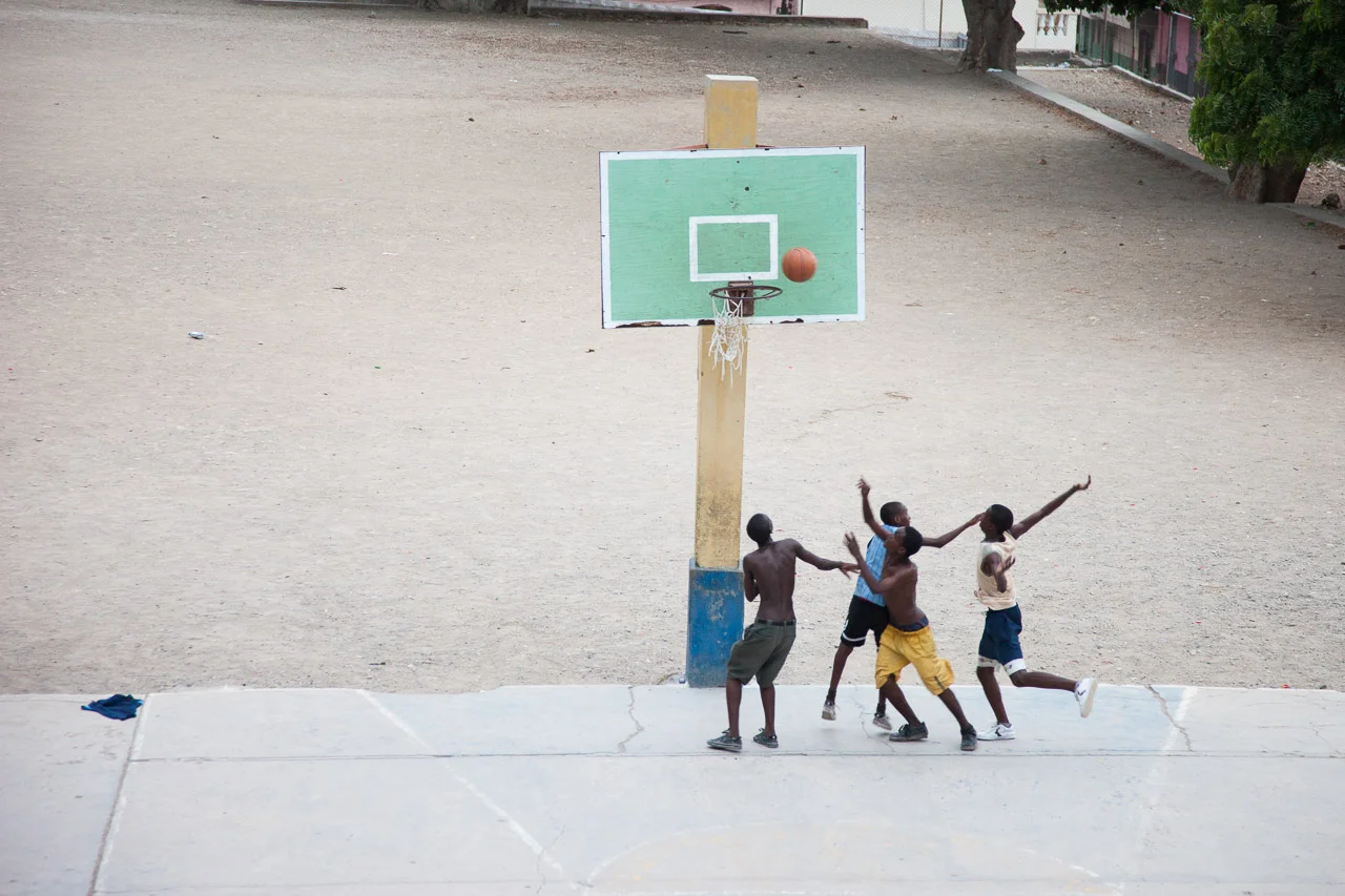 Four young boys playing basketball on an outdoor court, one boy appears to be taking a shot while the other three defend.