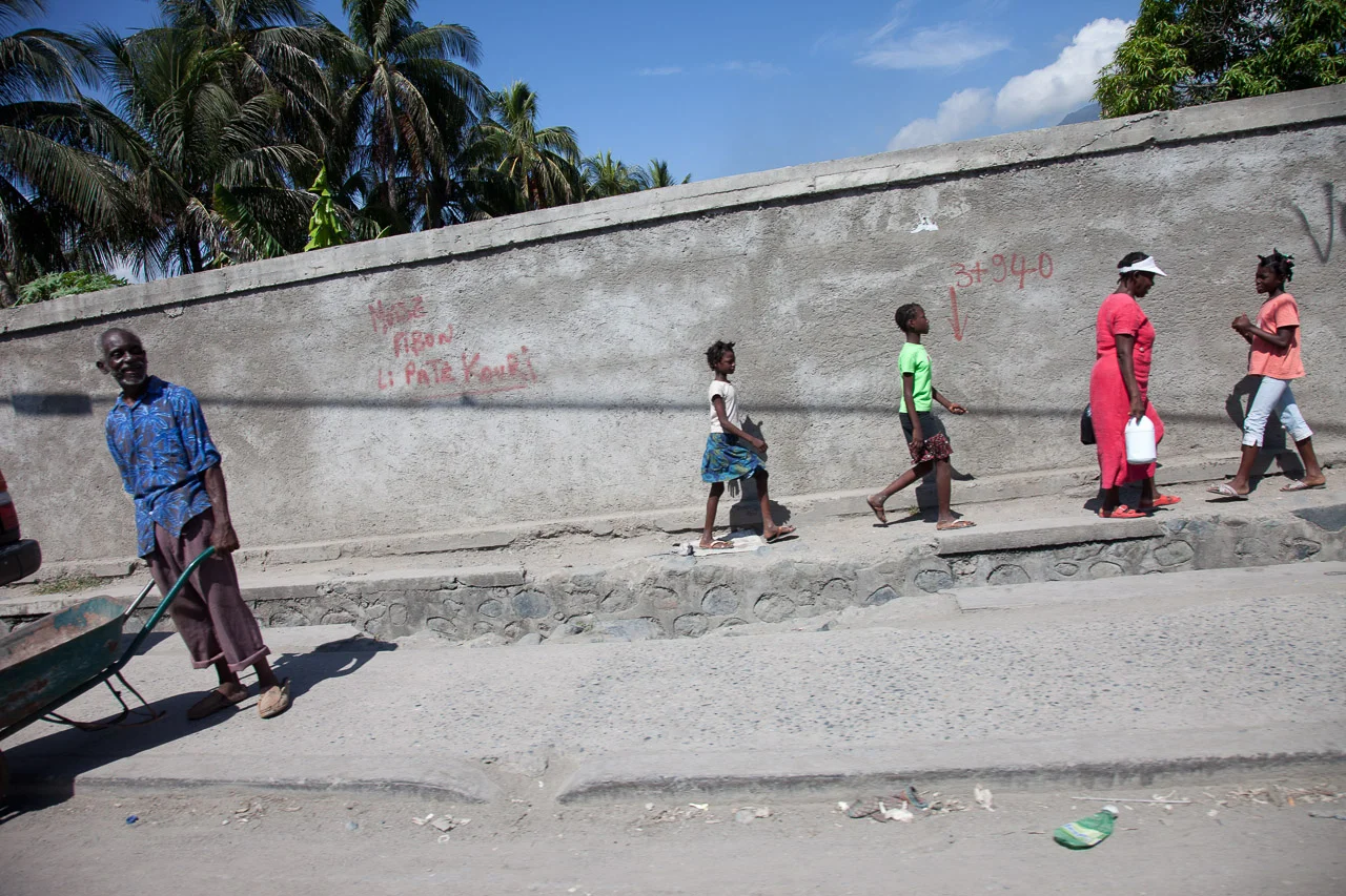 A man with a wheelbarrow walks past a group of children and an adult woman walking along a sidewalk beside a wall with graffiti. The background features palm trees and a blue sky.