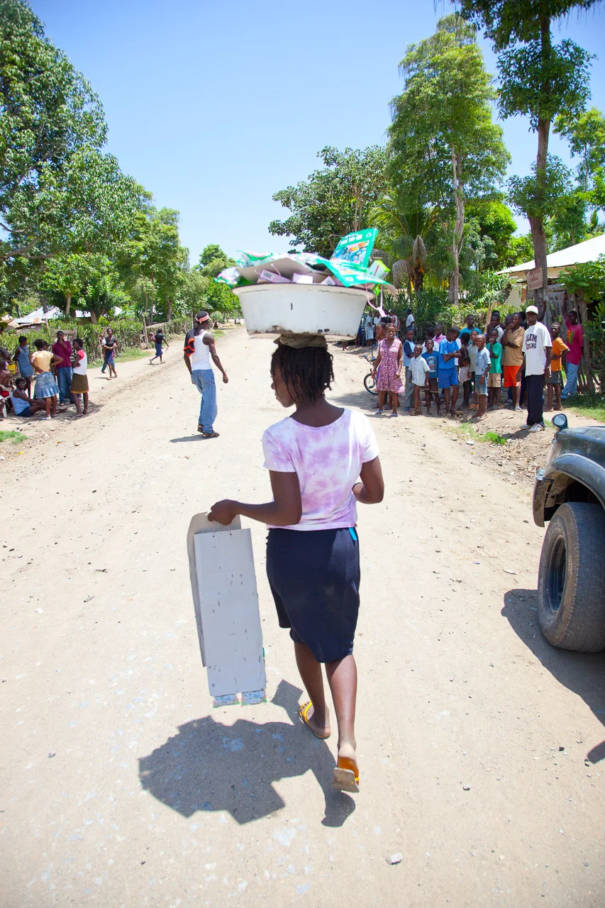 A young girl walking down a dirt street carrying a rectangular box with a bowl of other items on her head. Several children and adults are gathered on the sides of the street under sunny weather, with trees and small houses in the background.